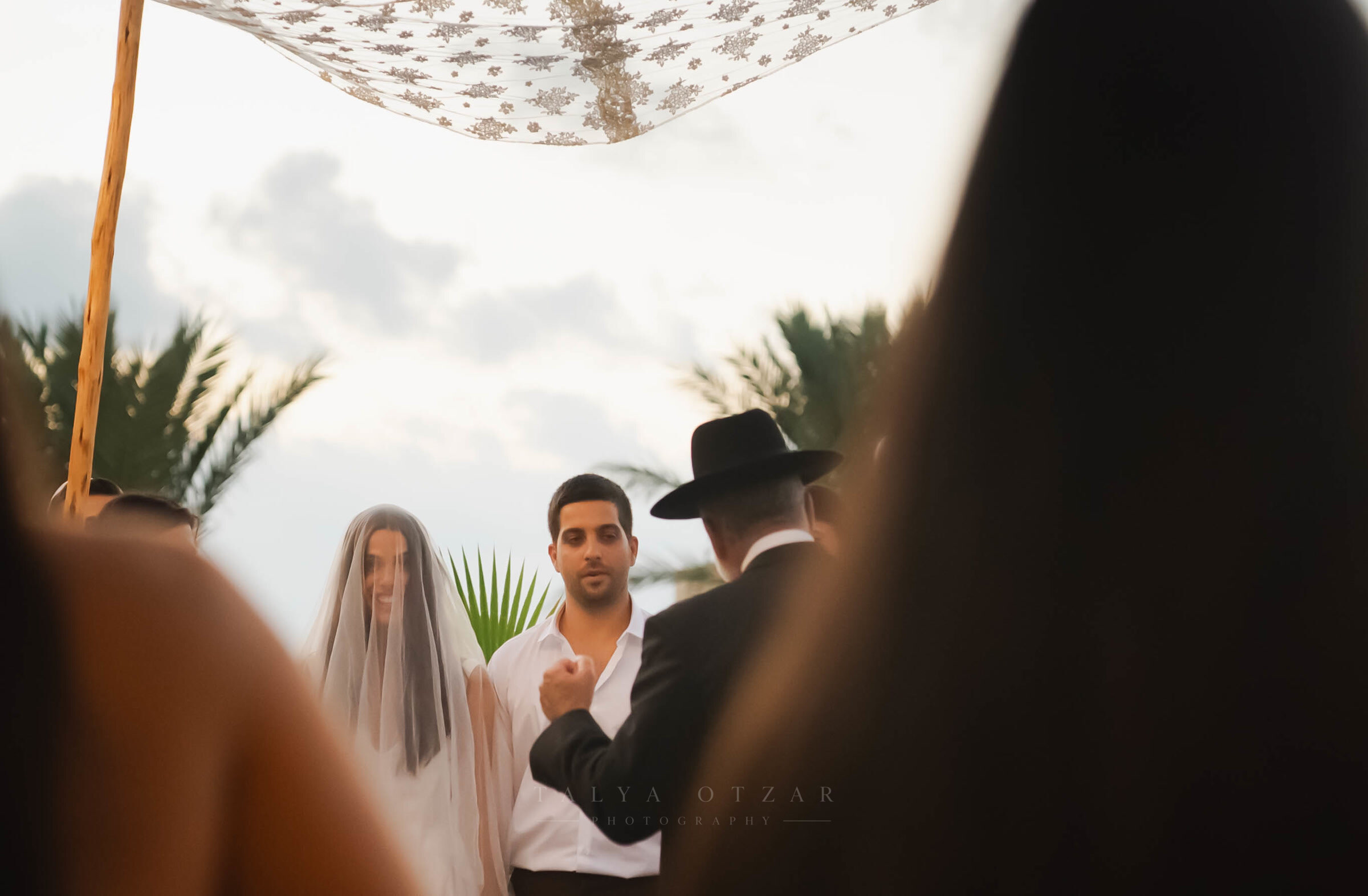 The bride and groom under the lace chuppah, seen between the shoulders of the guests
