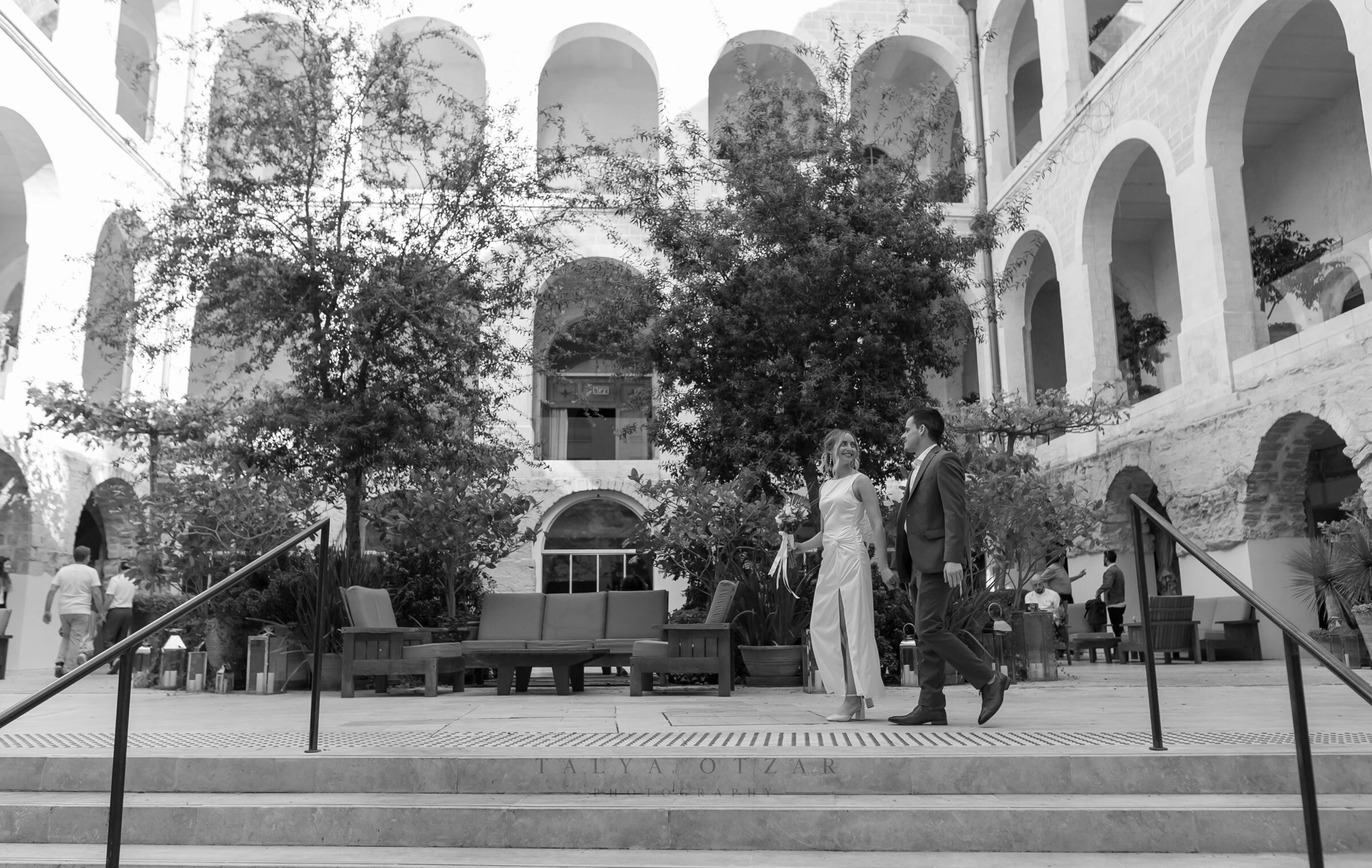 Bride and groom crossing the arcaded courtyard, the arches rising above them