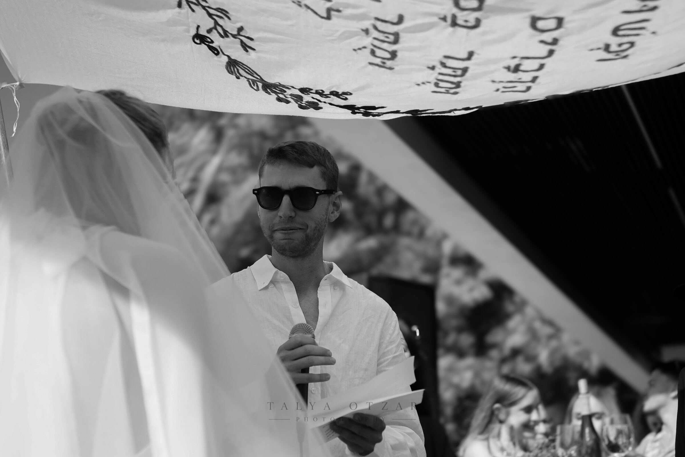 The groom reading aloud under the embroidered chuppah cloth, the bride's veil at the edge of the frame