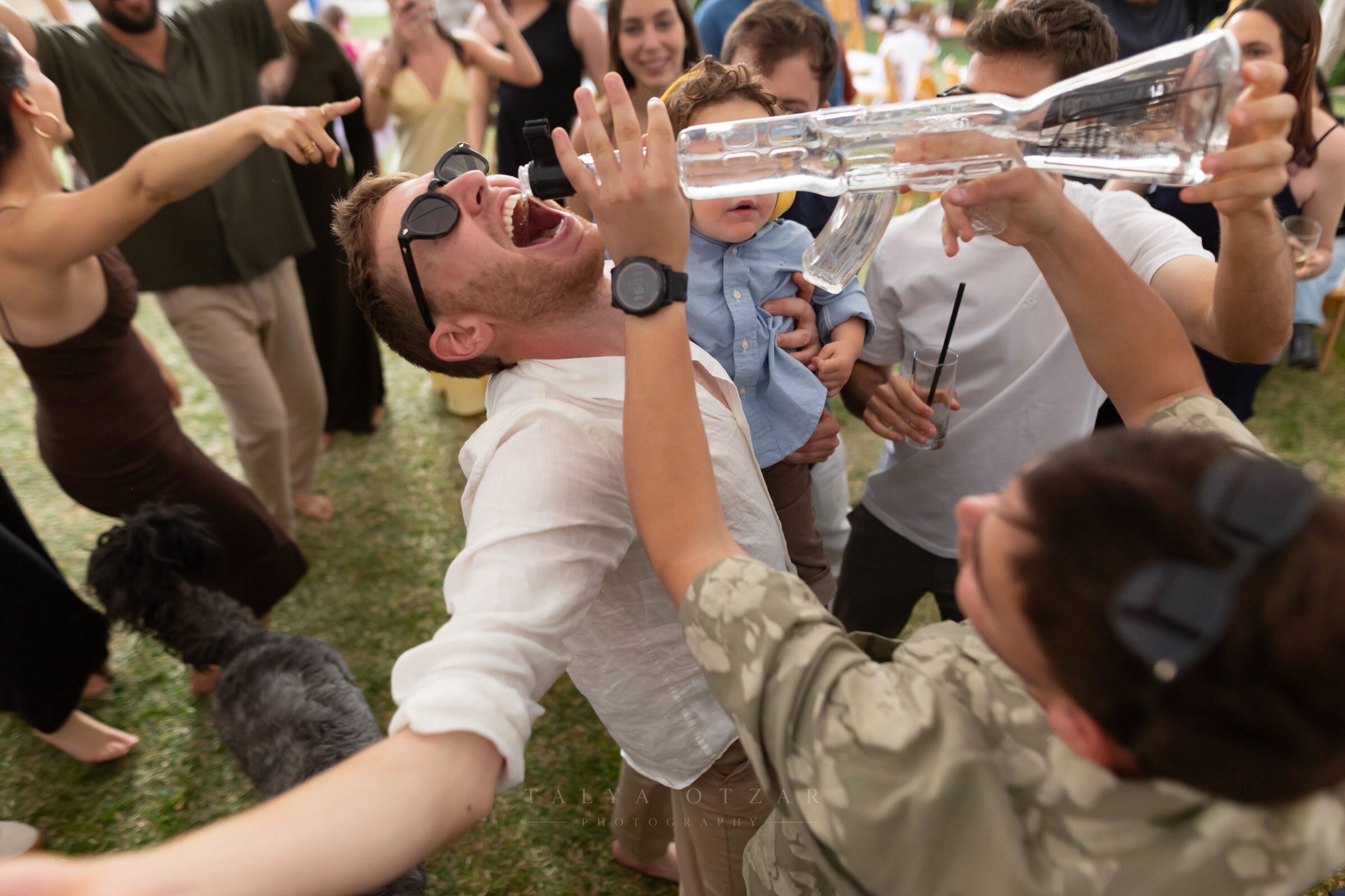 A guest laughing open-mouthed as friends pour a drink for him on the grass