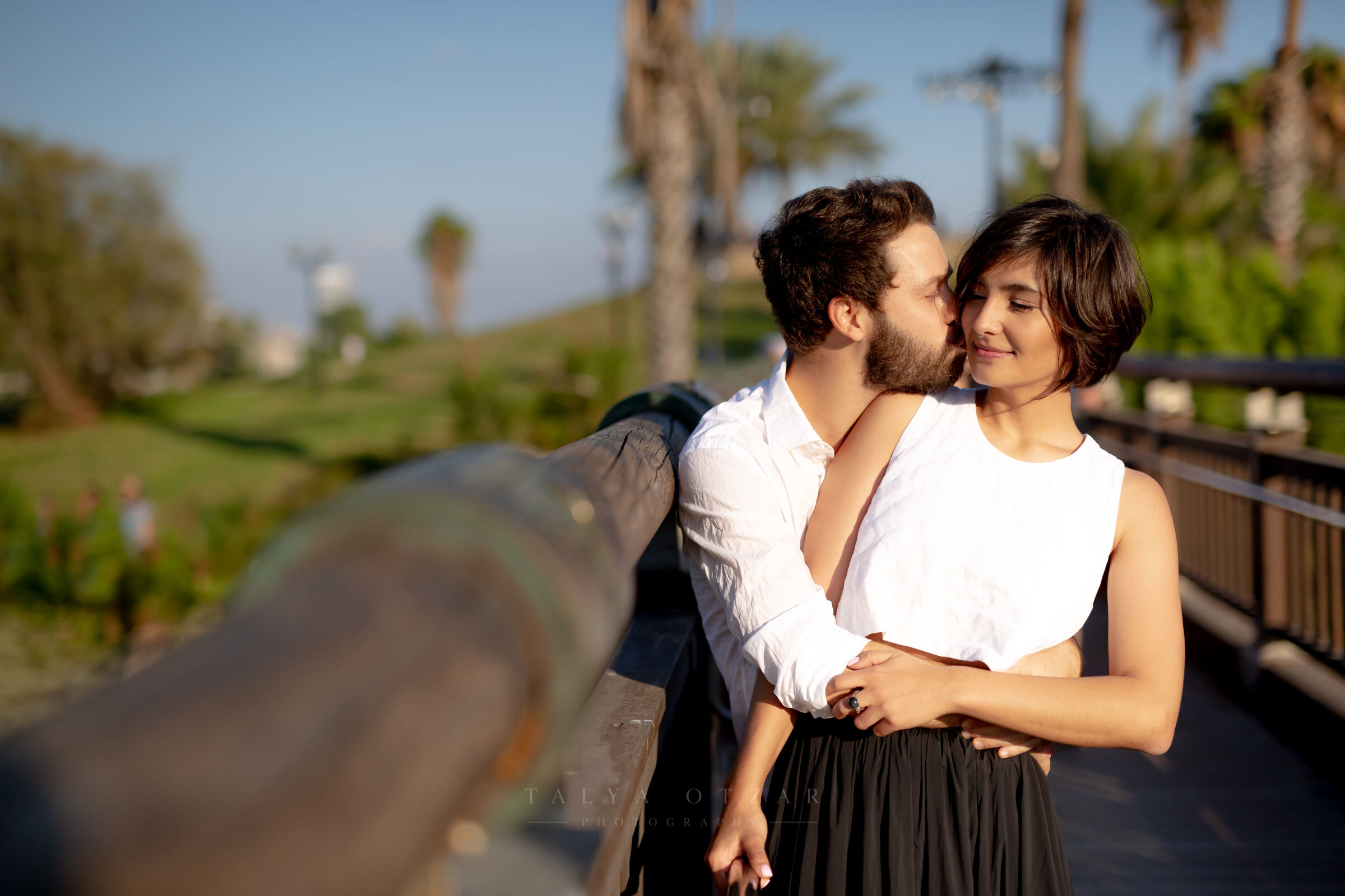 The couple at the seafront railing at golden hour, the groom kissing her cheek