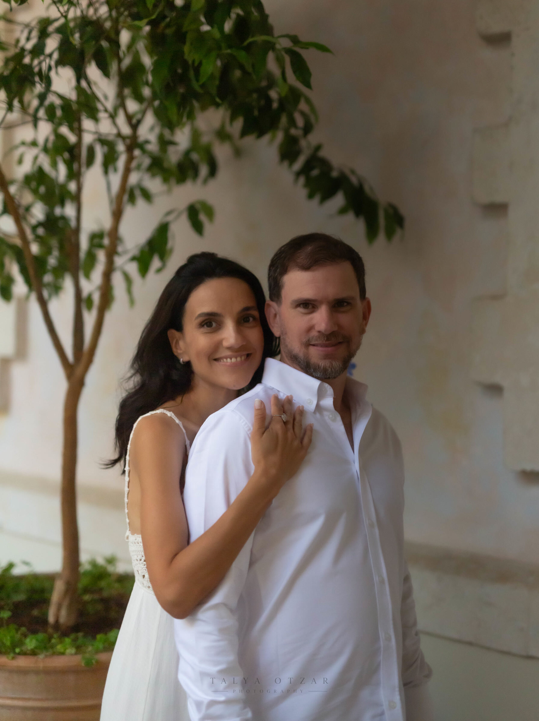 Couple portrait against a plaster wall, the bride's chin resting on the groom's shoulder