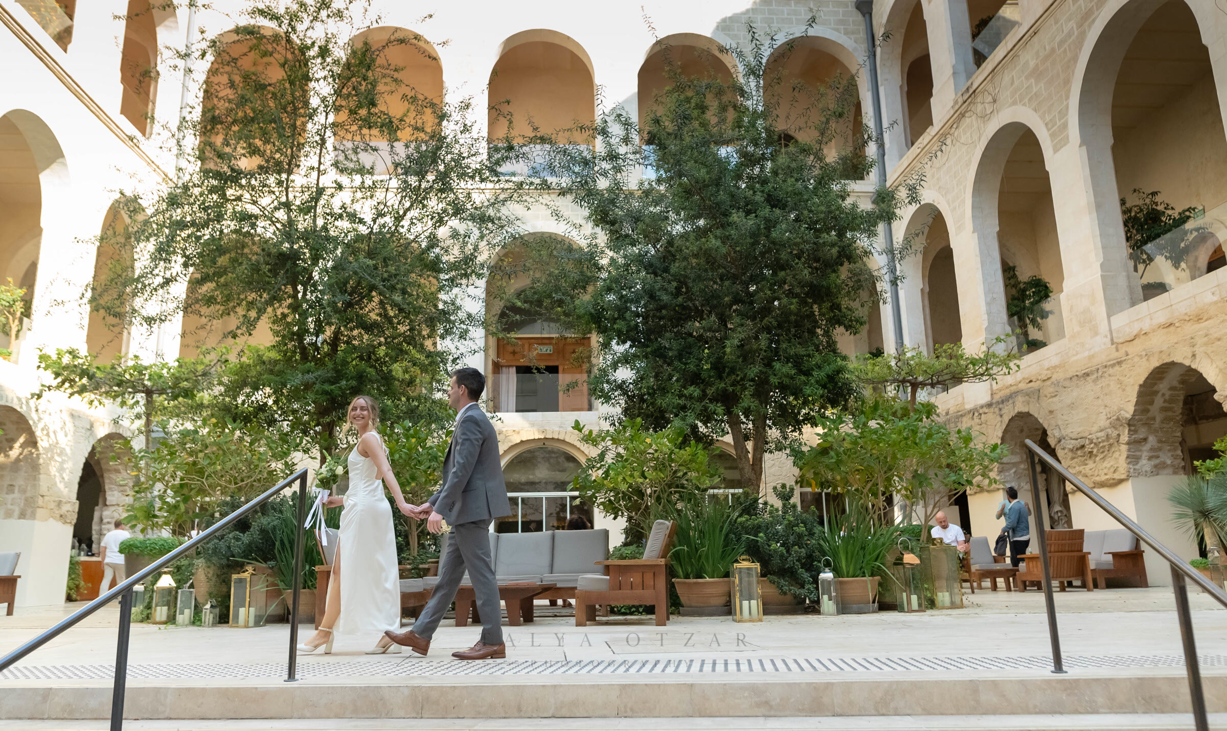 Bride and groom walking hand in hand across the arcaded stone courtyard