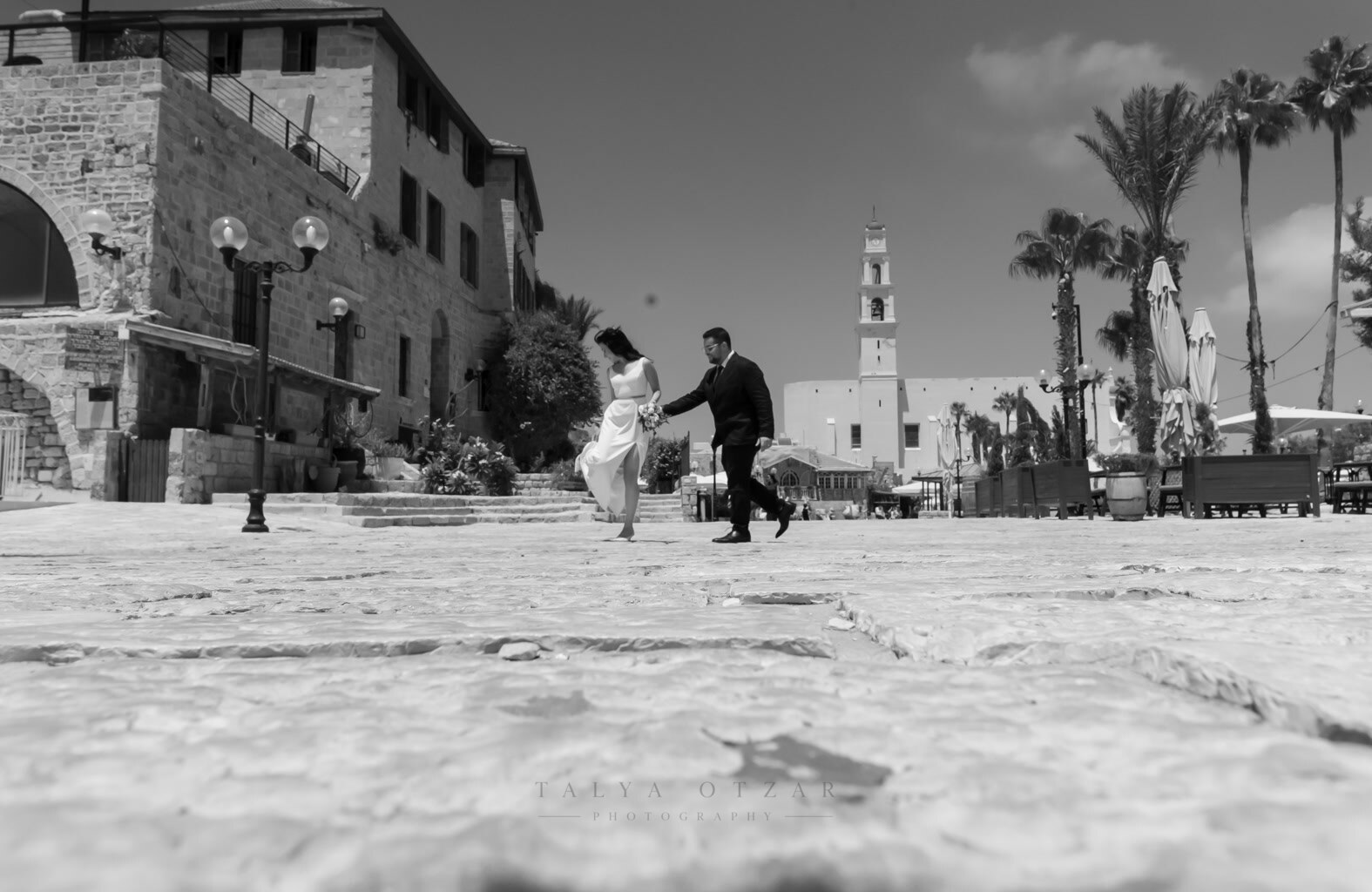 The couple crossing the wide square in Old Jaffa, the church tower and palms behind them