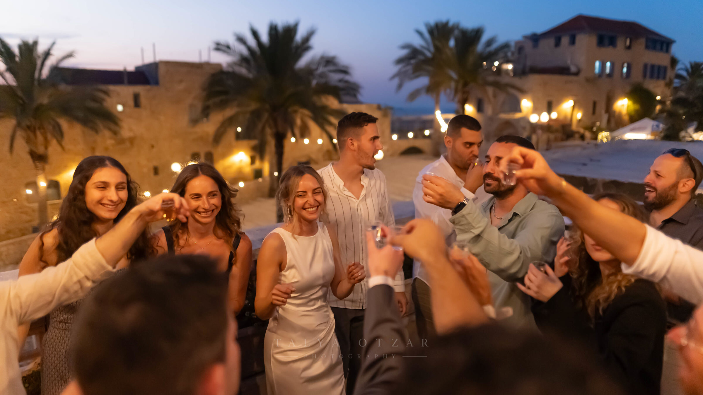 Guests raising a toast on the Jaffa rooftop at dusk, festoon lights and palms behind