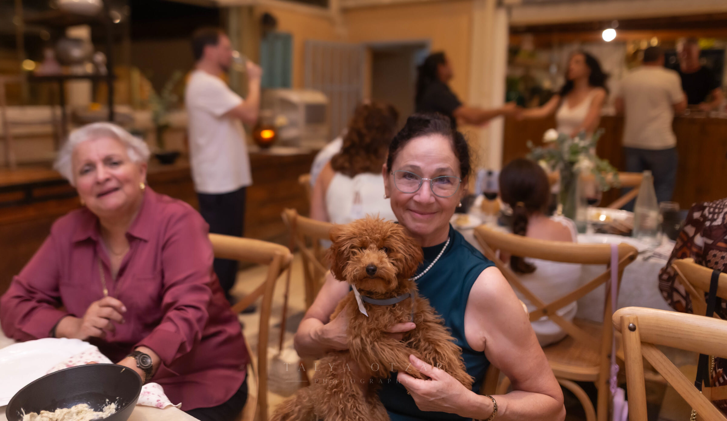 Two guests at the dinner table, one holding a small poodle on her lap