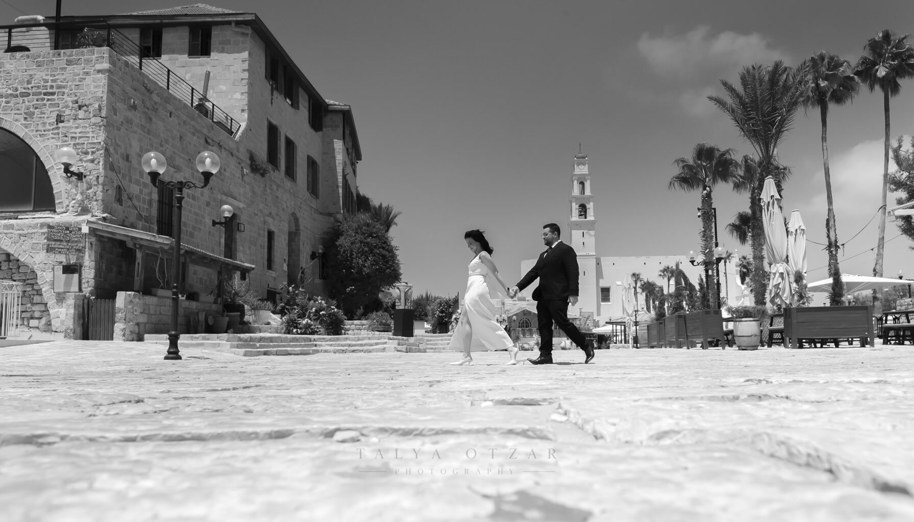 The couple crossing the Jaffa square hand in hand, the bell tower rising behind