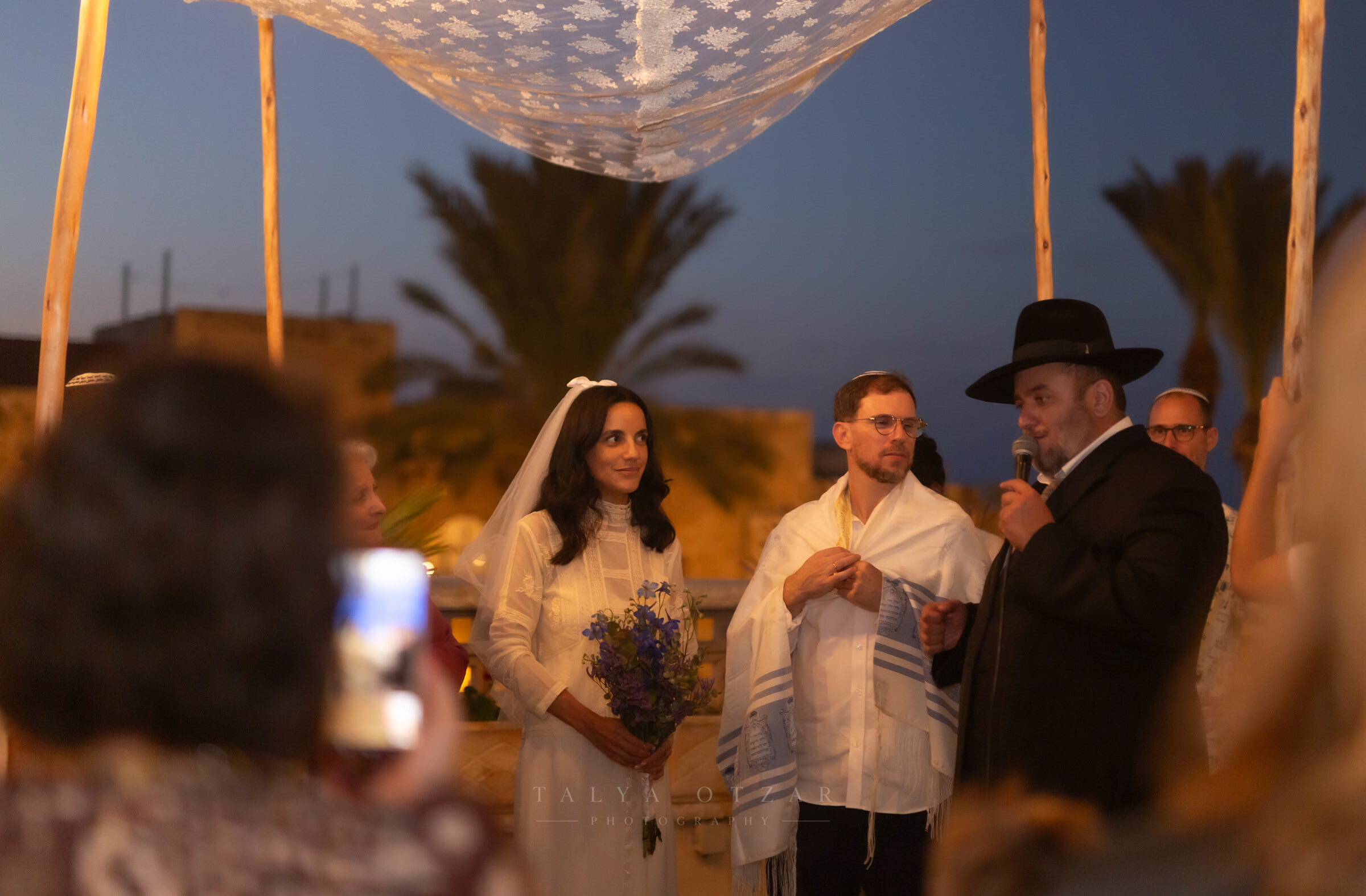 Bride and groom under the lace chuppah at dusk, the rabbi speaking and a guest filming from the front