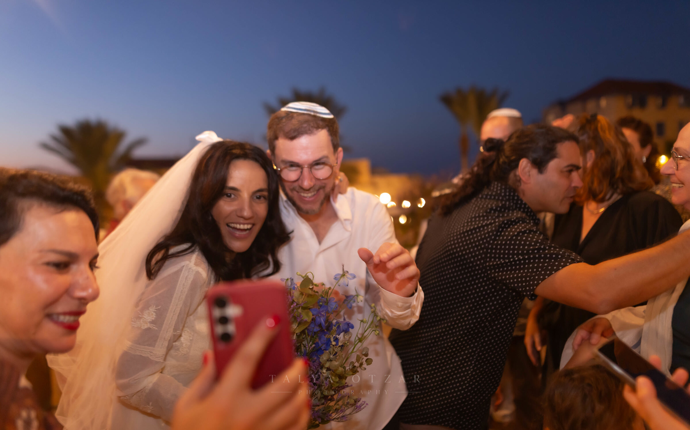 The couple laughing as a guest leans in for a photograph with them at dusk