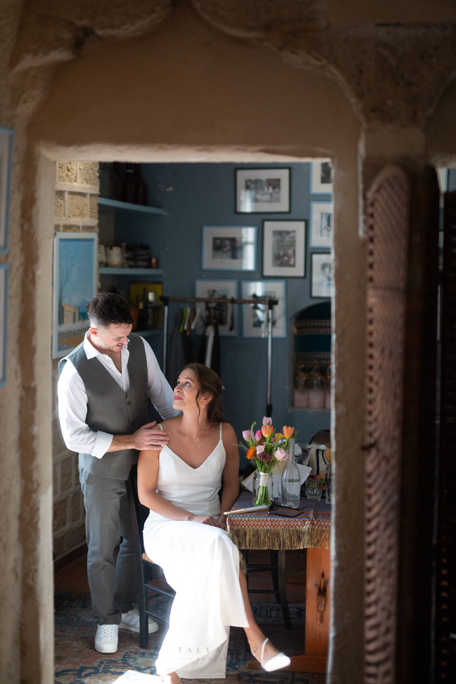 The couple in a blue-walled room hung with photographs, seen through a stone archway