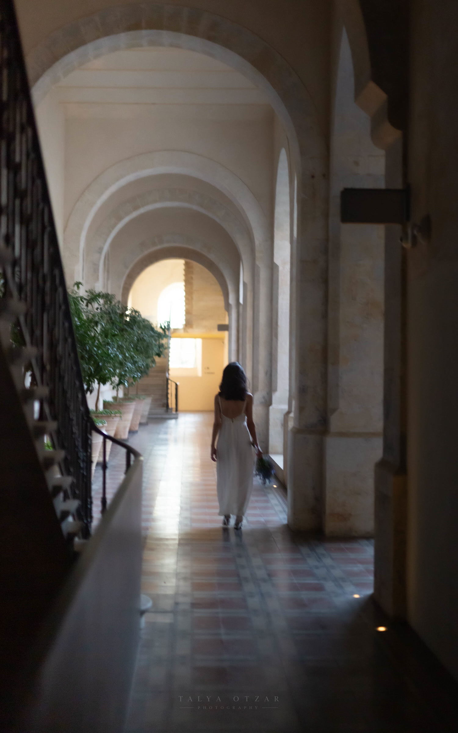 The bride walking away down a long arcaded corridor lined with potted plants