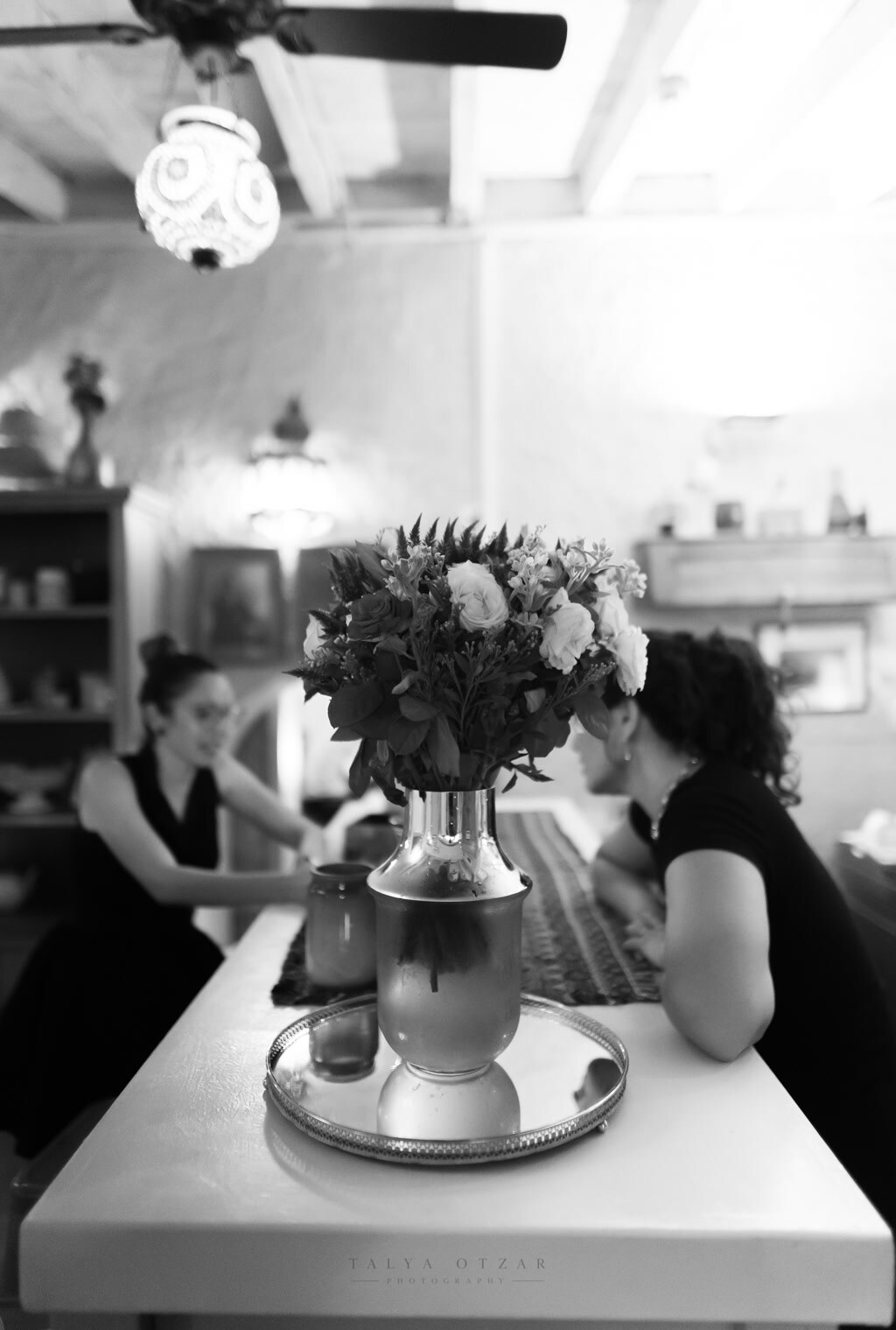 A vase of roses on a silver tray, two guests talking beyond it