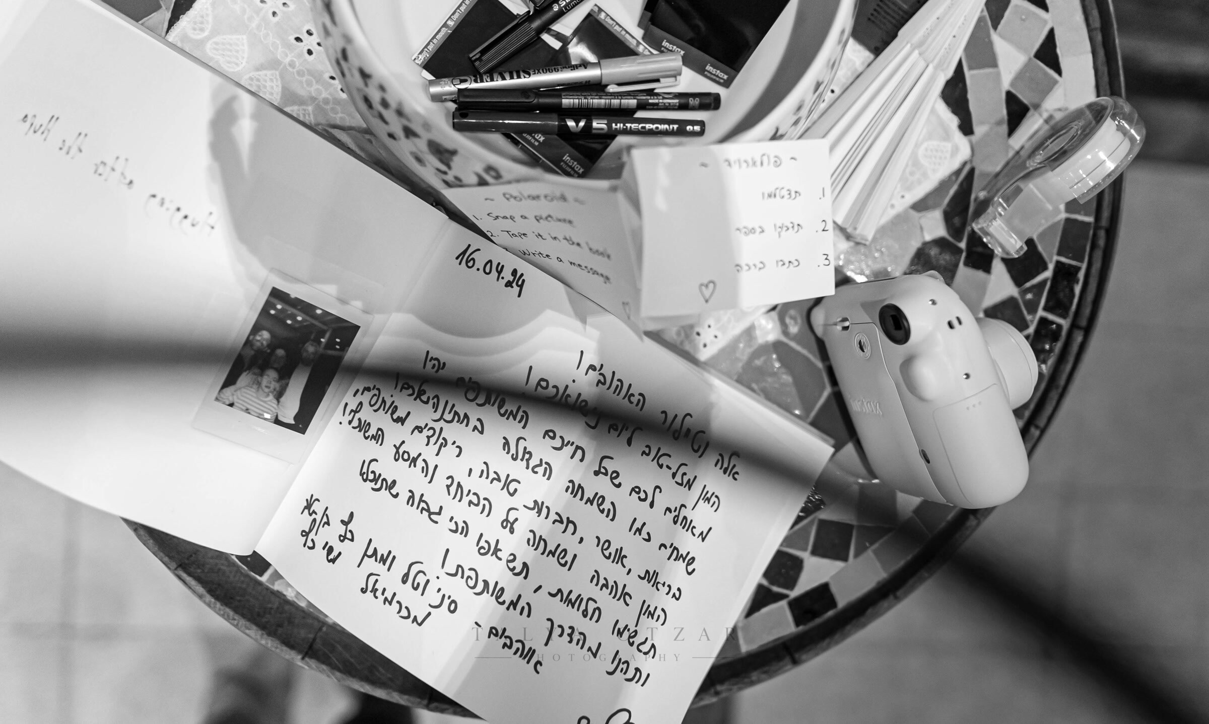 Handwritten vows and notes in Hebrew spread across a mosaic table beside marker pens and an instant camera