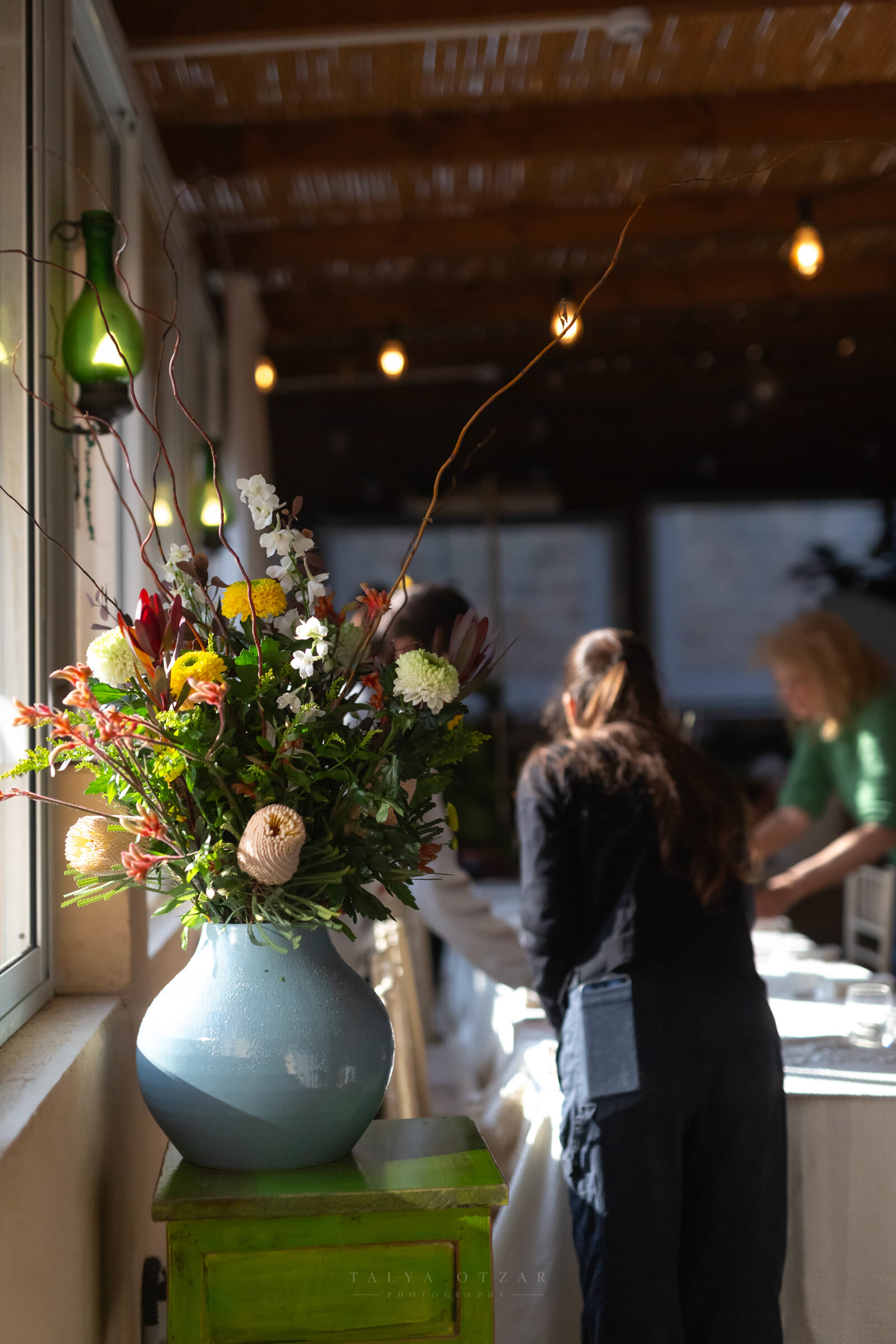 A tall arrangement of native flowers in a blue jug while the tables are laid behind