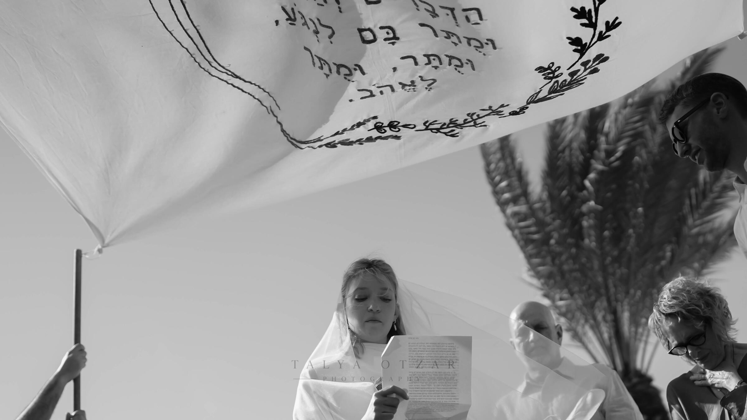 The bride reading aloud beneath the chuppah cloth embroidered in Hebrew