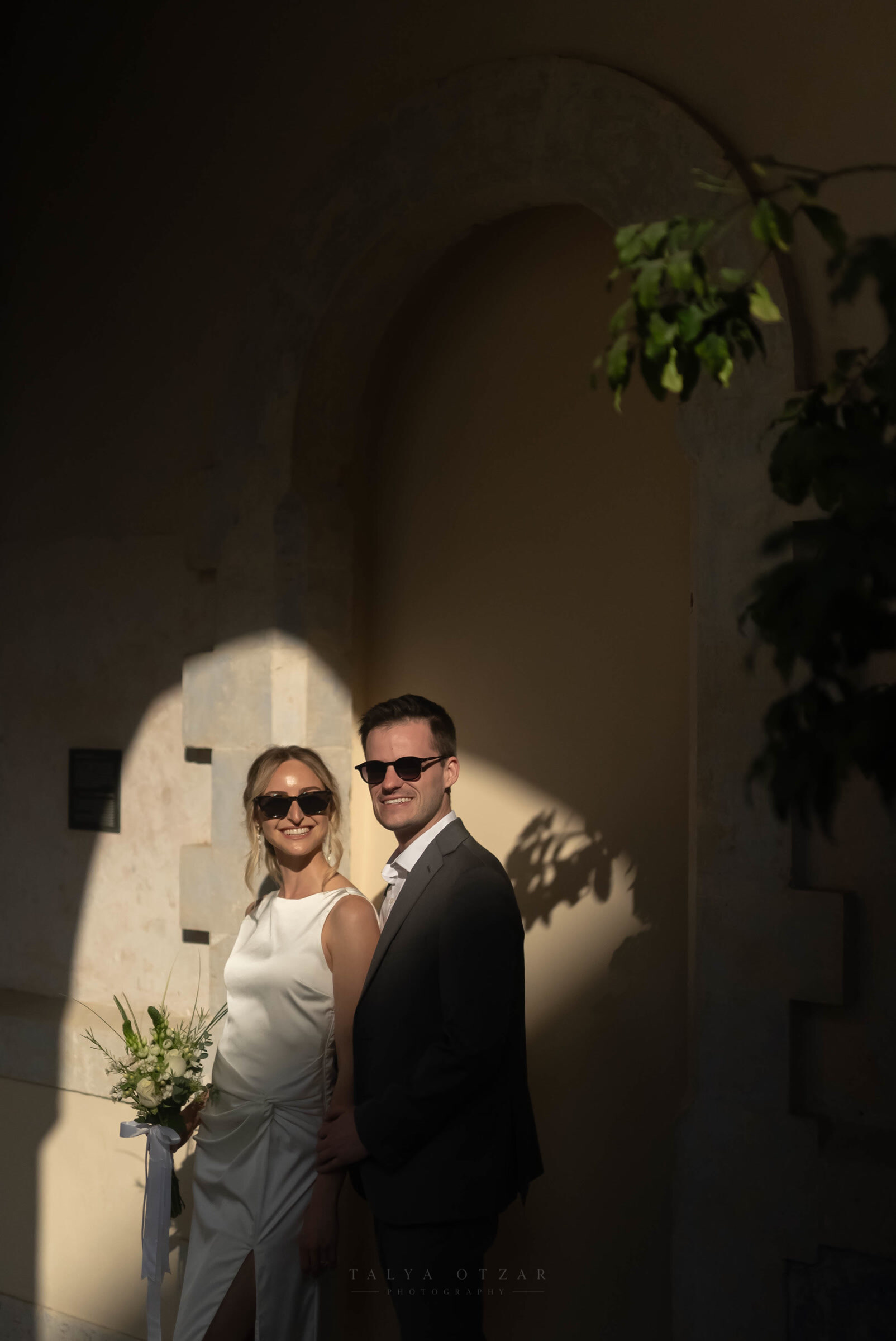 Bride and groom standing in a pool of low sun against an arched stone wall