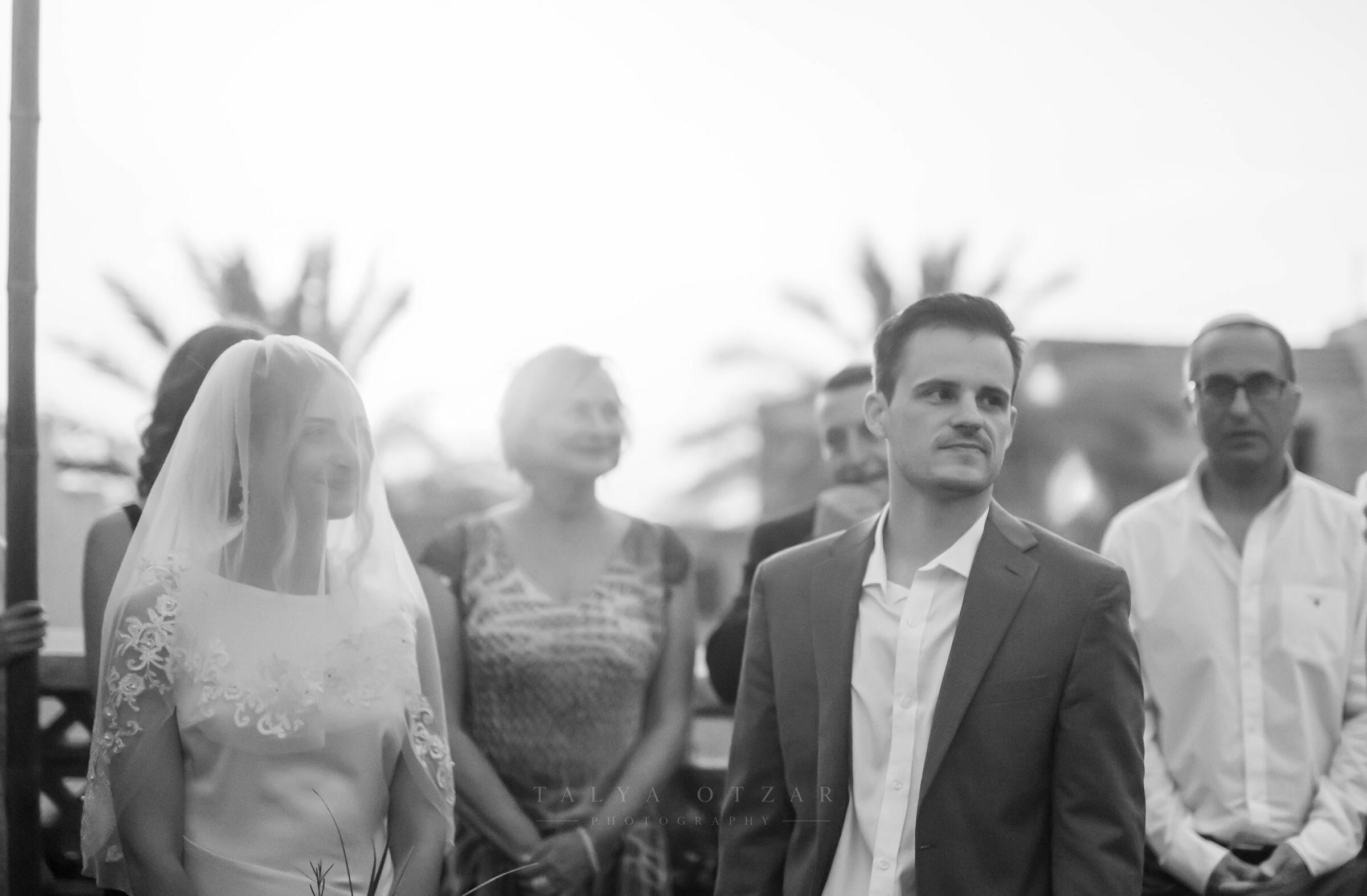 Bride and groom standing at the ceremony, guests and palm trees behind them