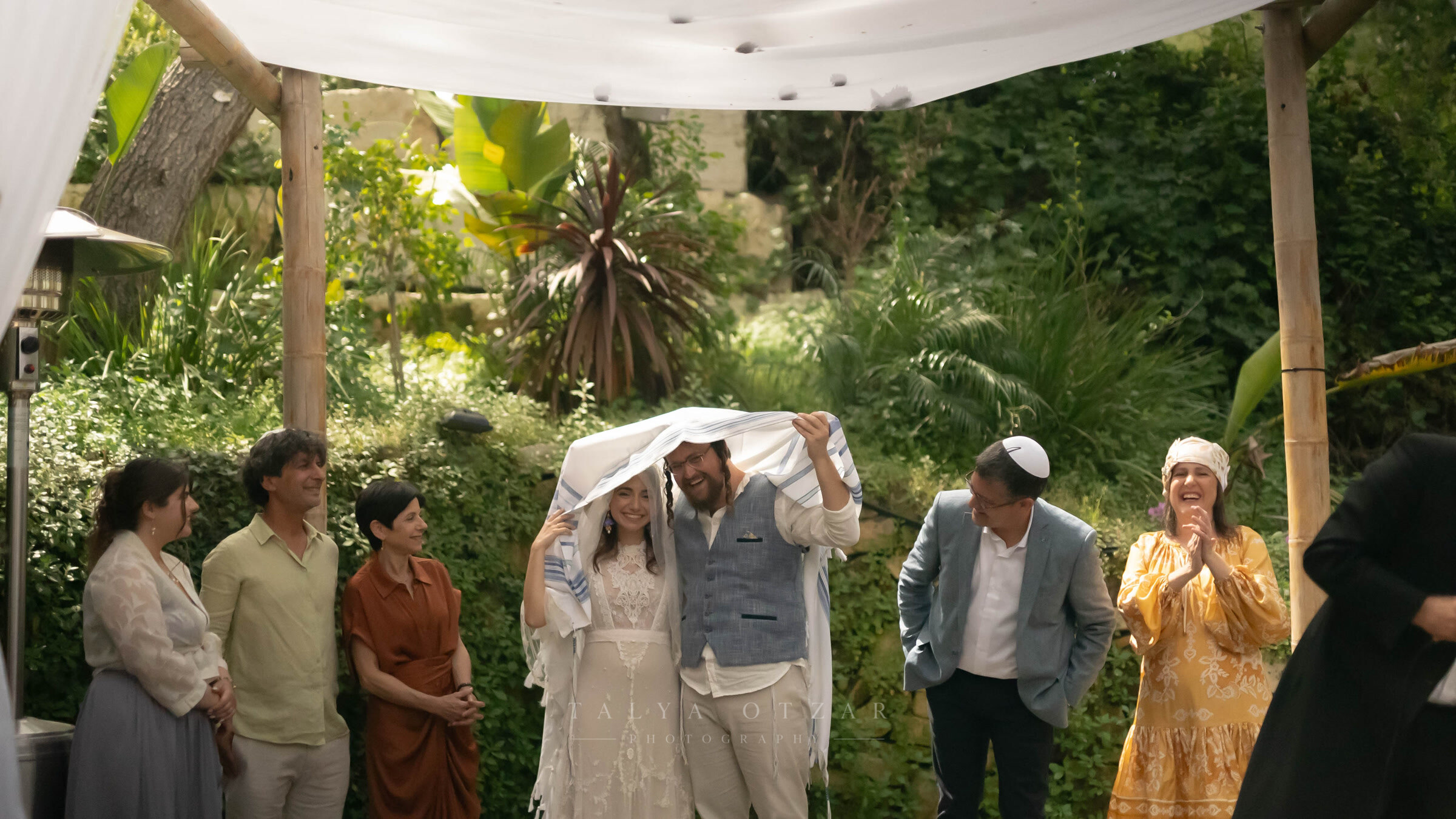 The couple under a tallit held above their heads in the garden, guests smiling around them