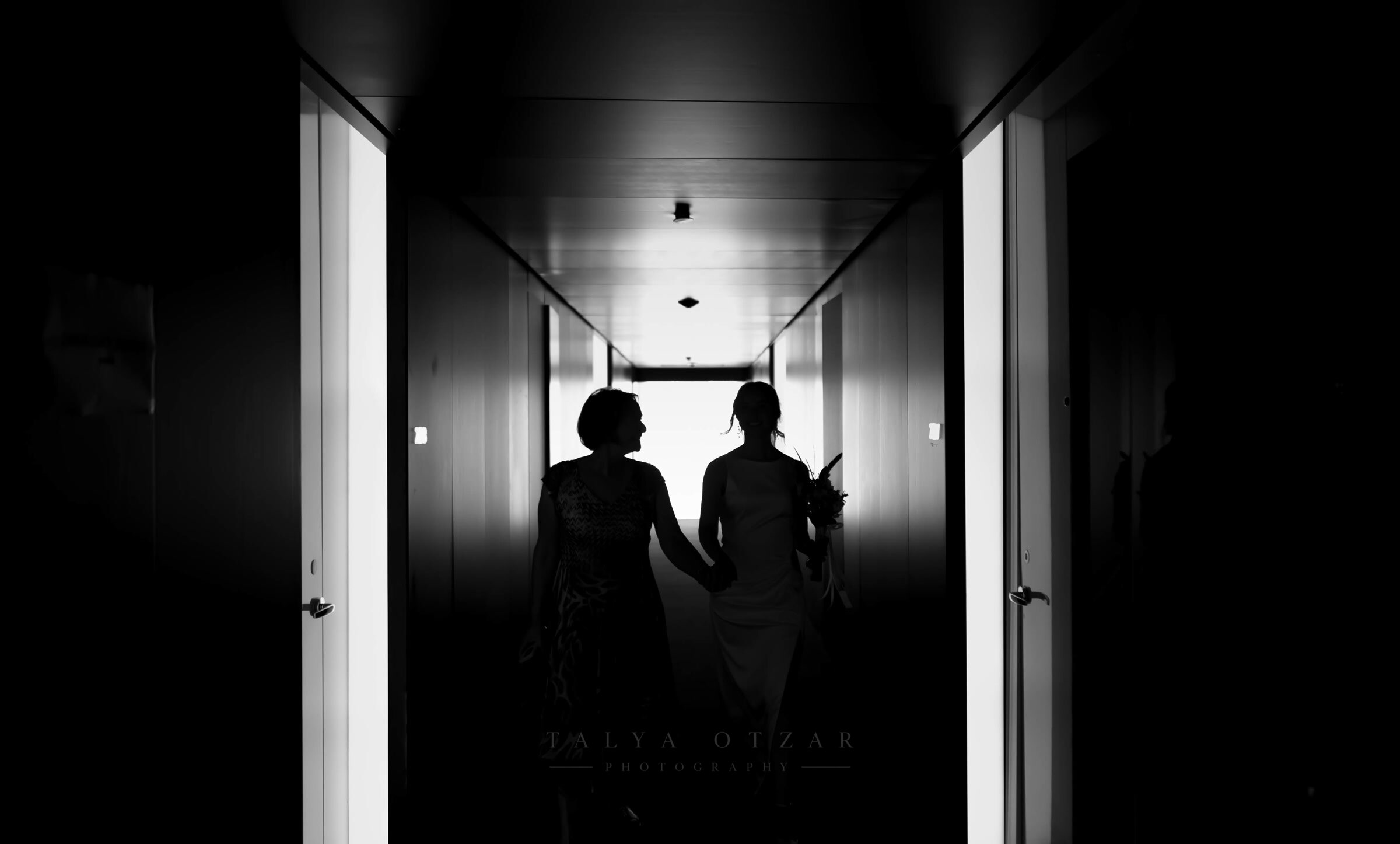 Bride and mother holding hands in silhouette down the length of a corridor