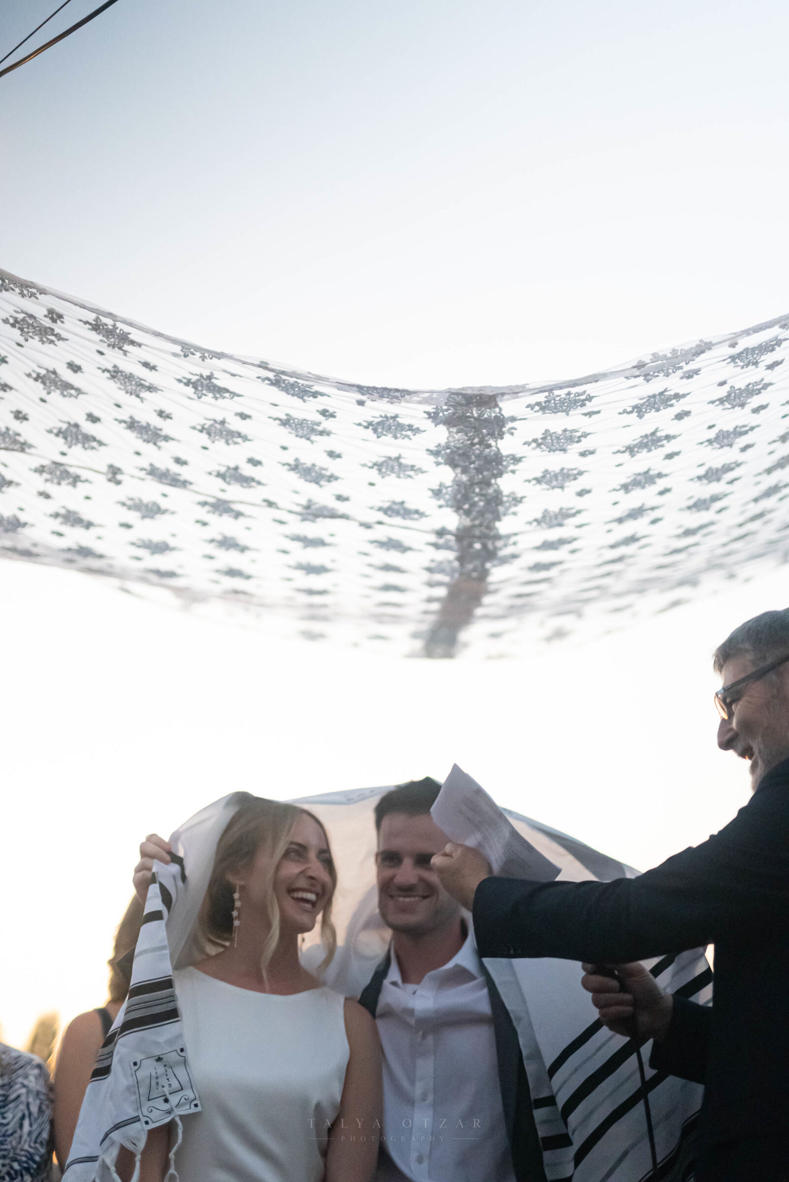 Bride and groom laughing under the lace chuppah as the officiant holds up the page