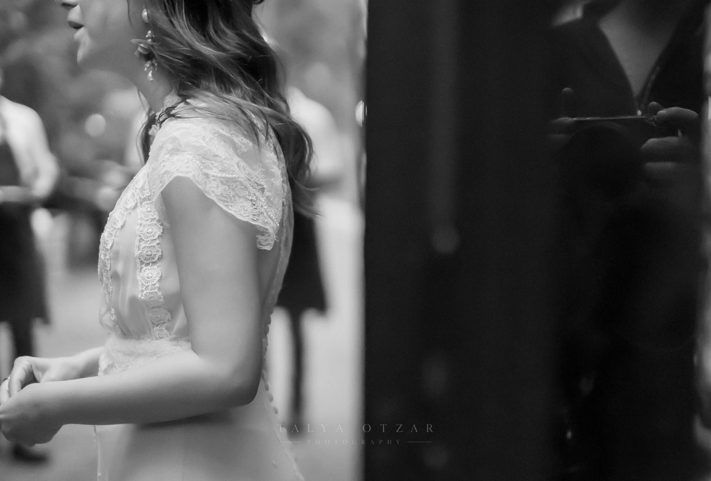 The bride in profile in her lace dress while a guest photographs her on a phone