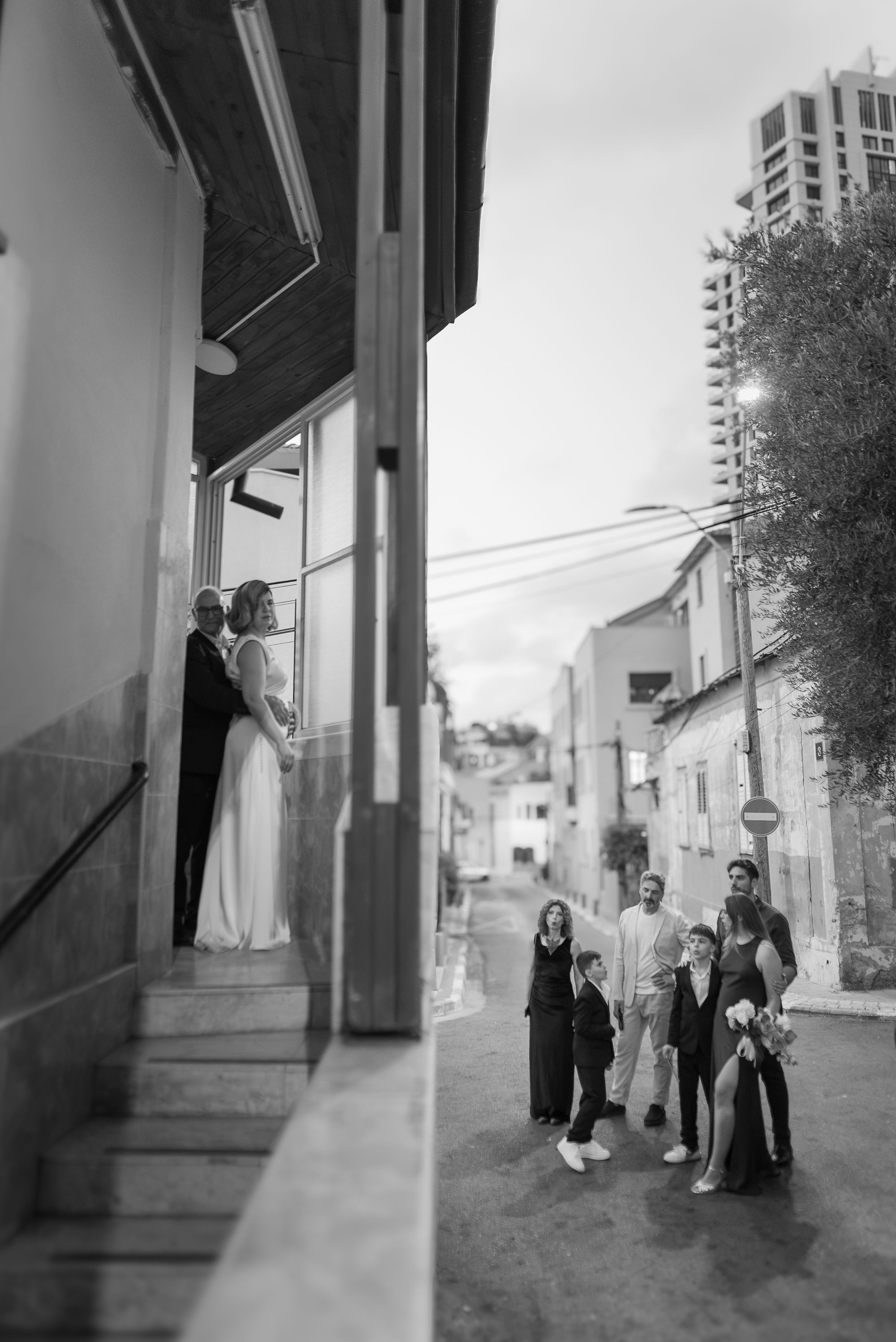The couple embracing at the top of the steps as their family waits in the street below, black and white