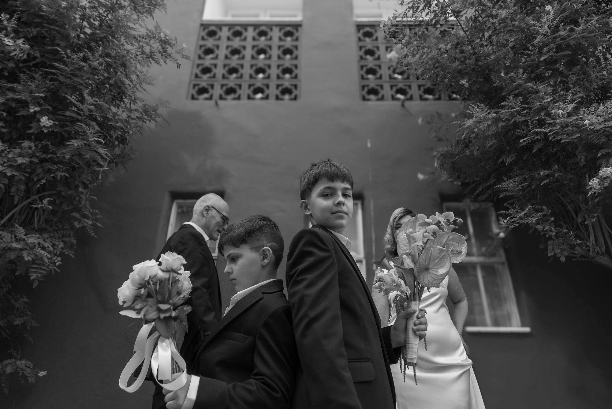 The couple and the two boys holding bouquets beneath a leafy wall, black and white