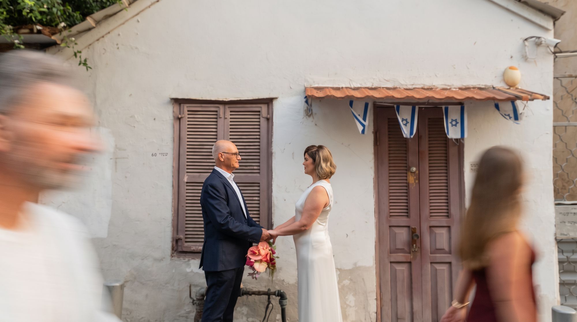 The couple holding hands at the ceremony outside a whitewashed house as guests pass in a blur