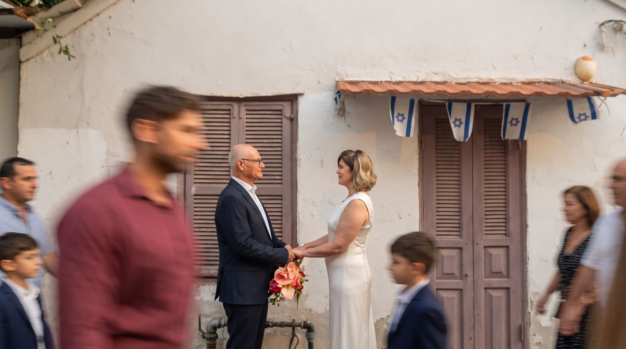 The bride and groom holding hands outside a whitewashed house as guests walk past in a blur
