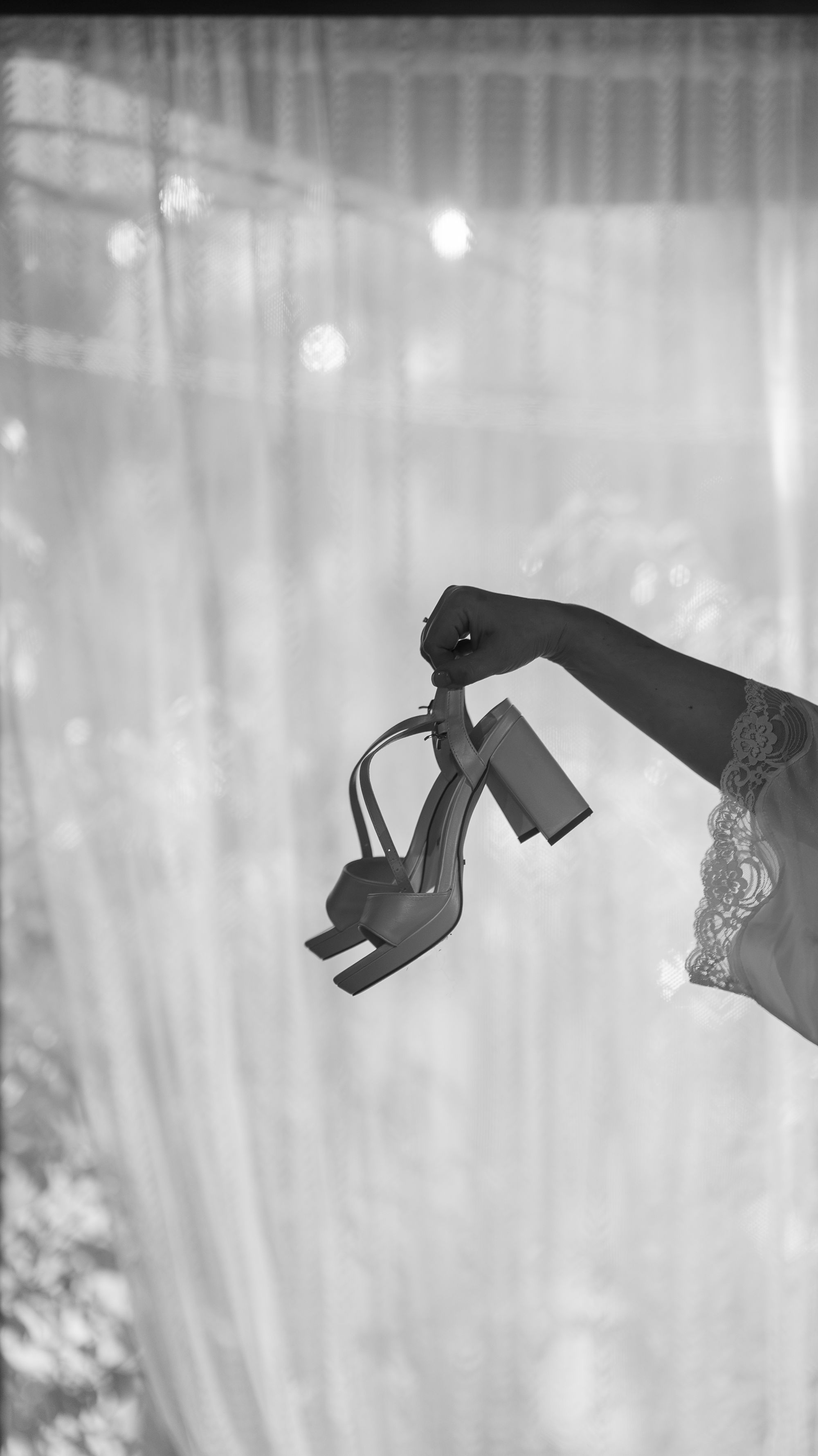 The bride's hand holding her sandals against a sheer curtain, black and white