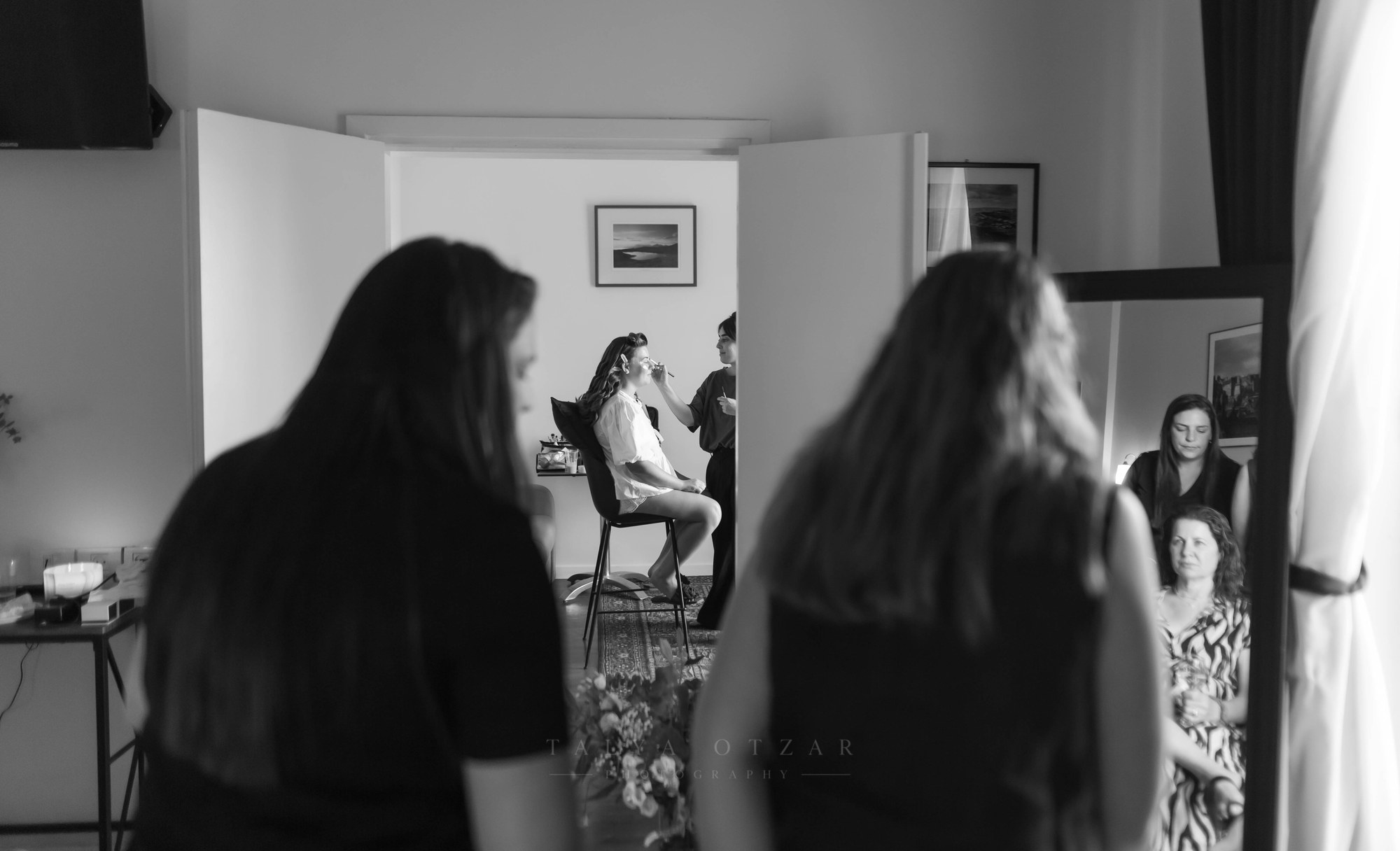 Friends watching the bride's make-up, reflected in a tall mirror, black and white