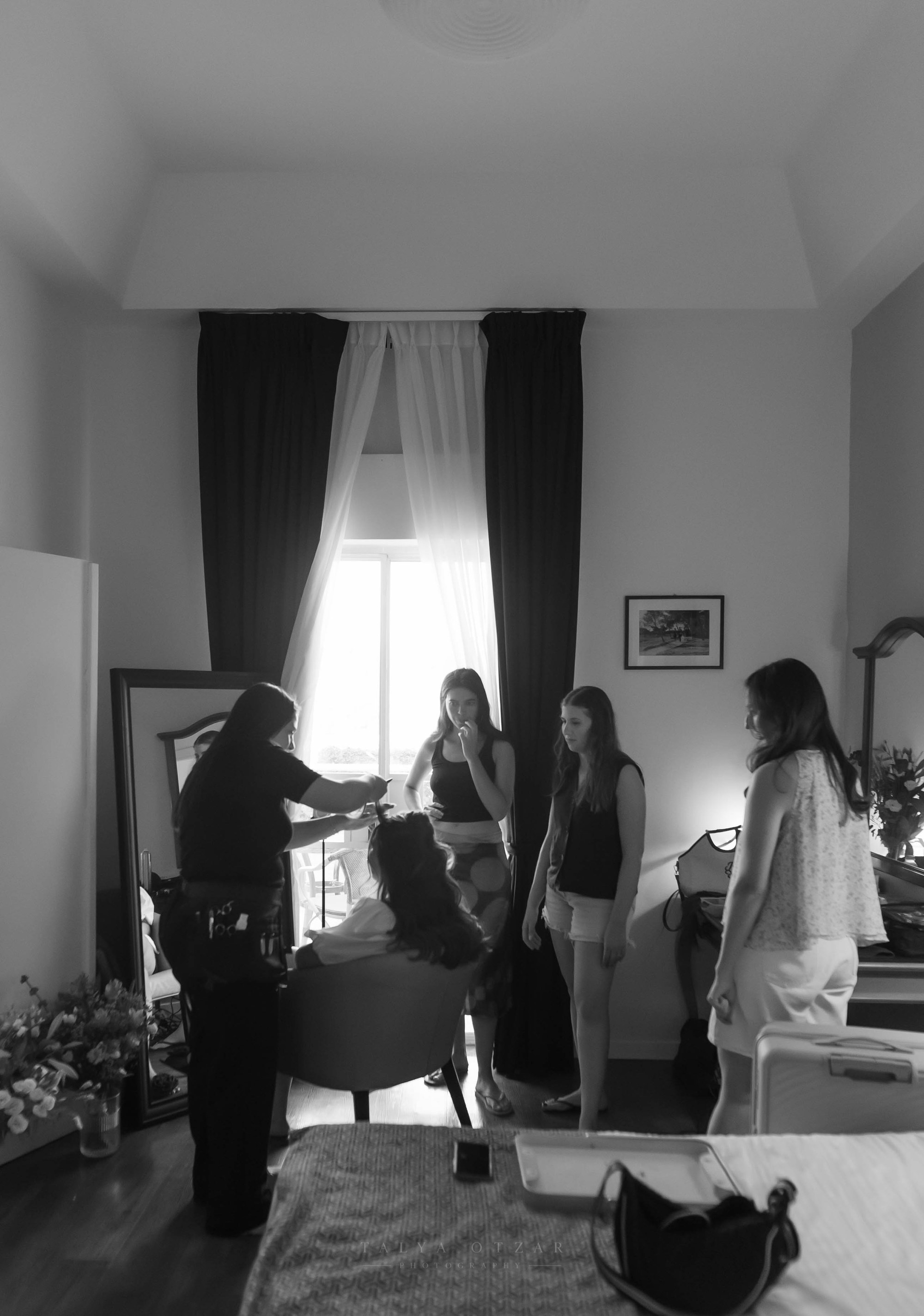 The bride's hair being styled by the window as her friends look on, black and white