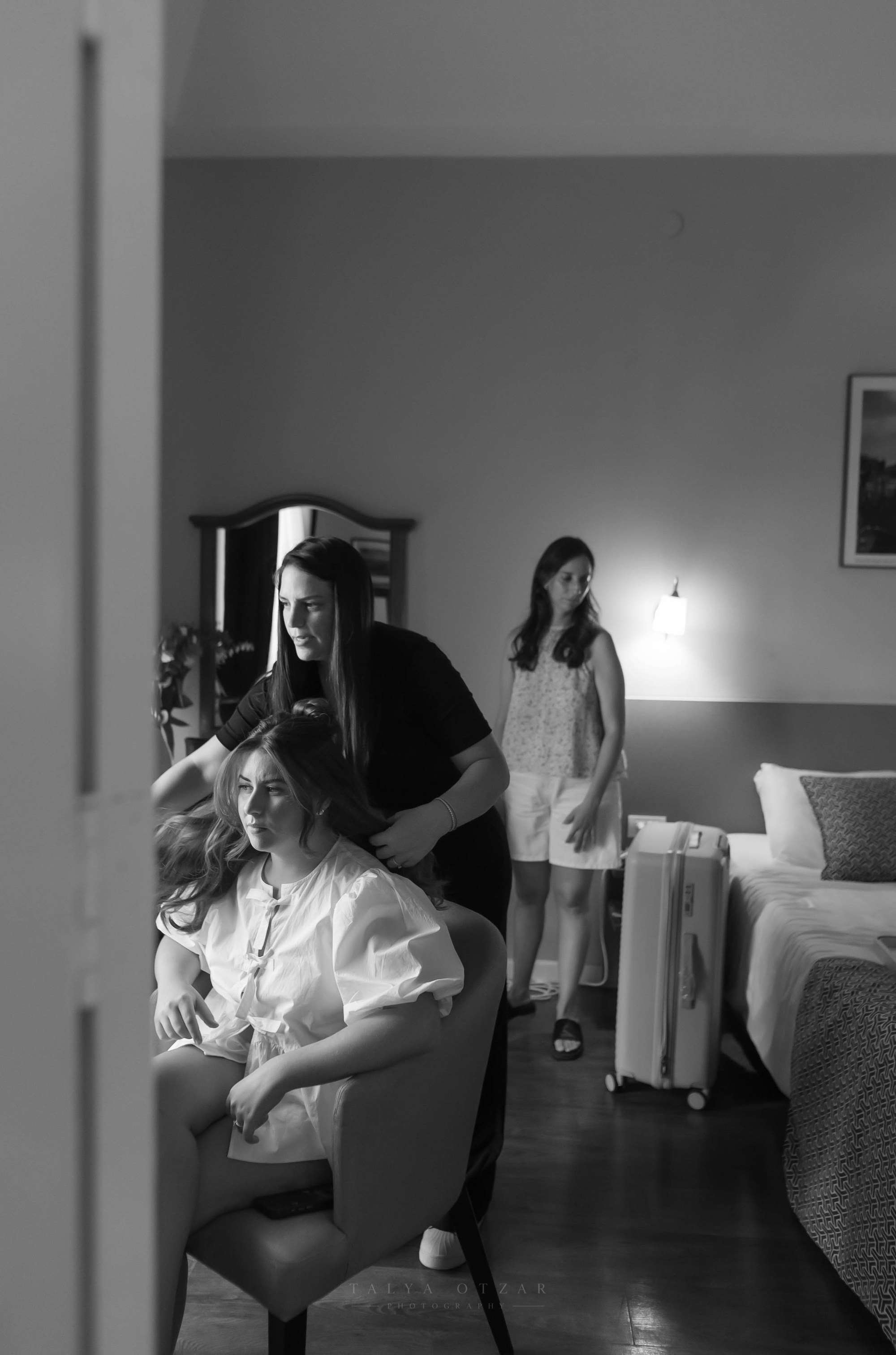 The bride in her robe as her hair is styled, a friend standing nearby, black and white