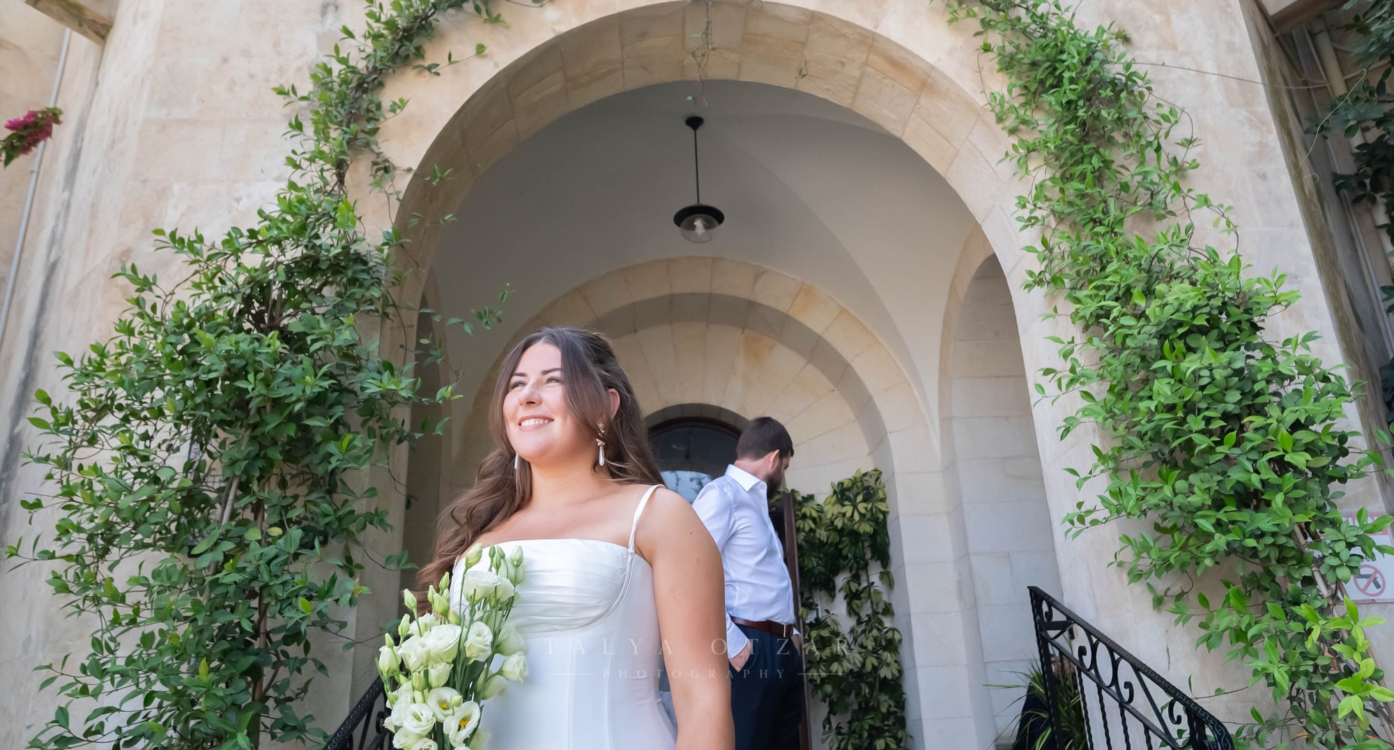The bride smiling beneath a stone arch hung with vines, the groom waiting behind her
