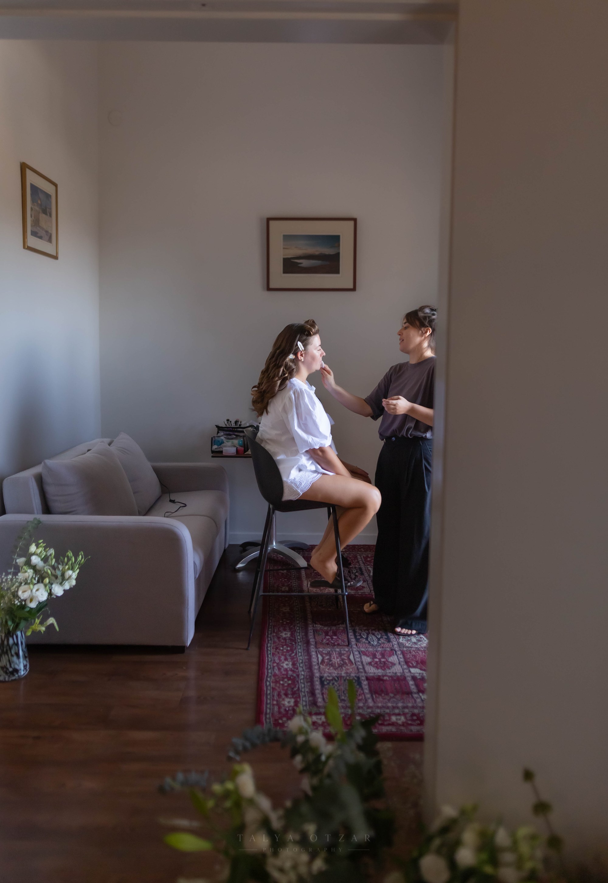 The bride having her make-up done, seen through a doorway past a vase of flowers