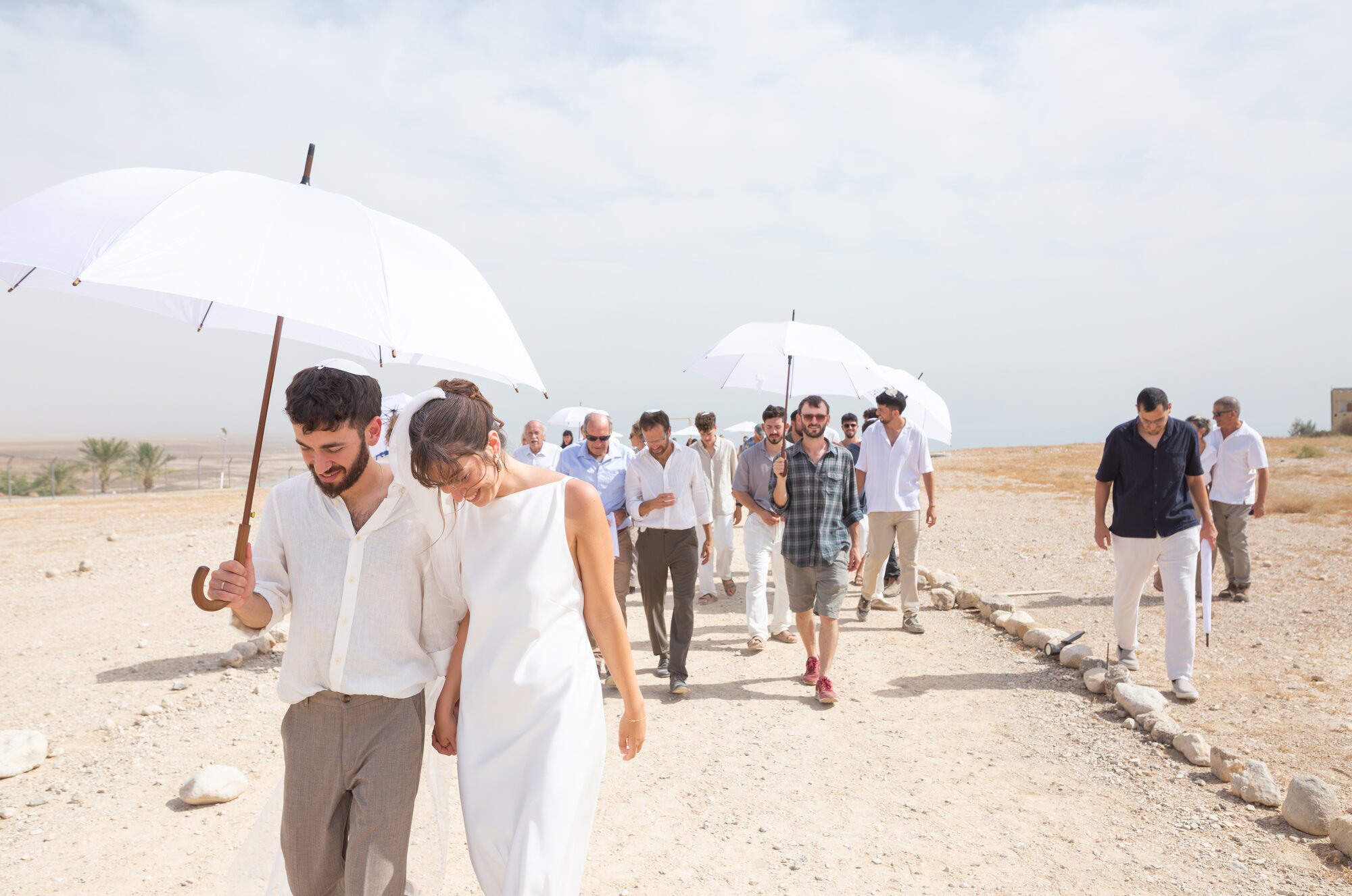 The couple walking along a desert path with their guests under white umbrellas