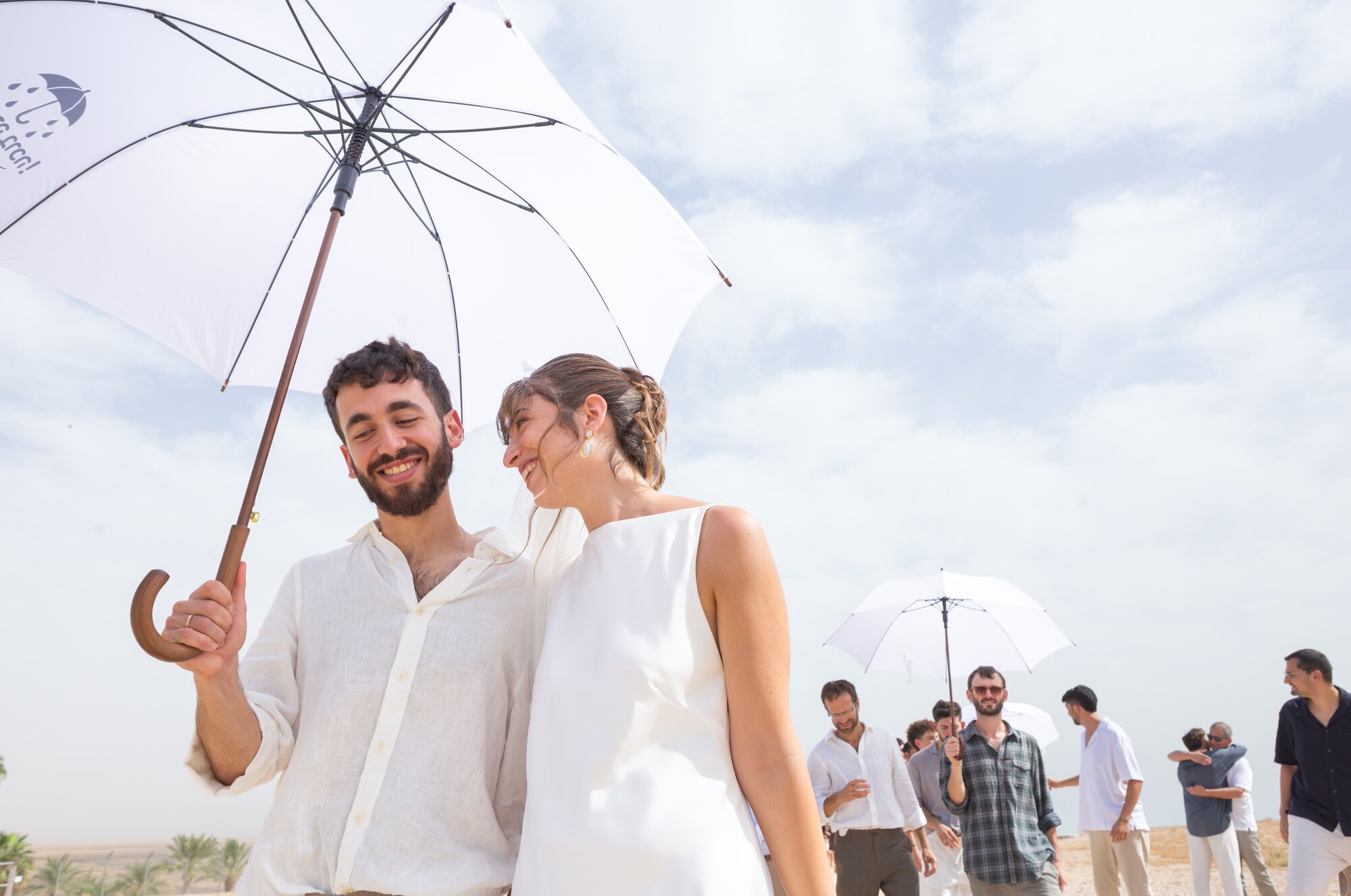The couple laughing under a white umbrella, friends following behind
