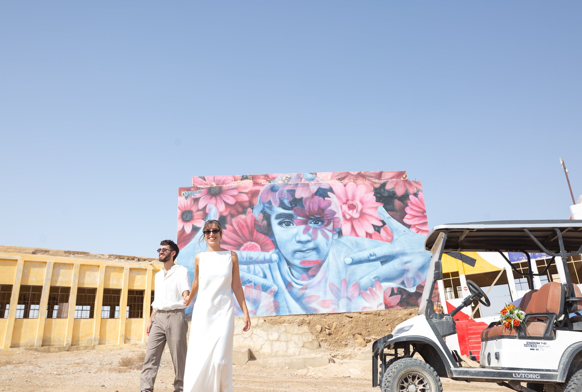 The couple walking past a giant flower mural, a golf cart parked beside them