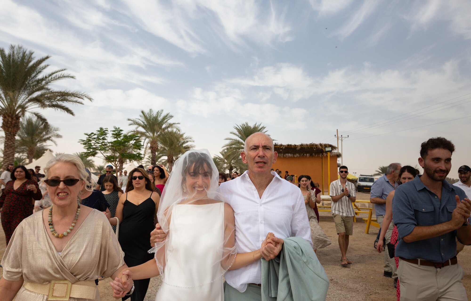 The veiled bride walking between her parents, surrounded by dancing guests