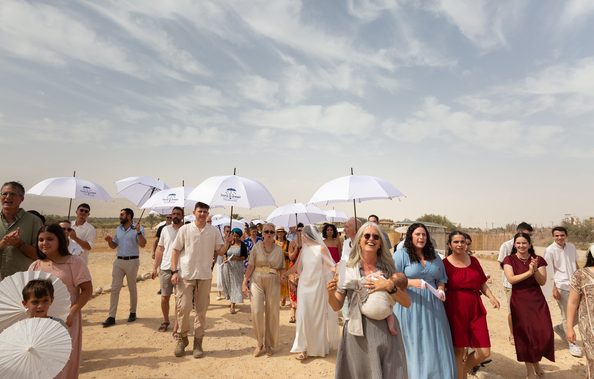 The procession of guests and white umbrellas crossing the desert under streaked clouds
