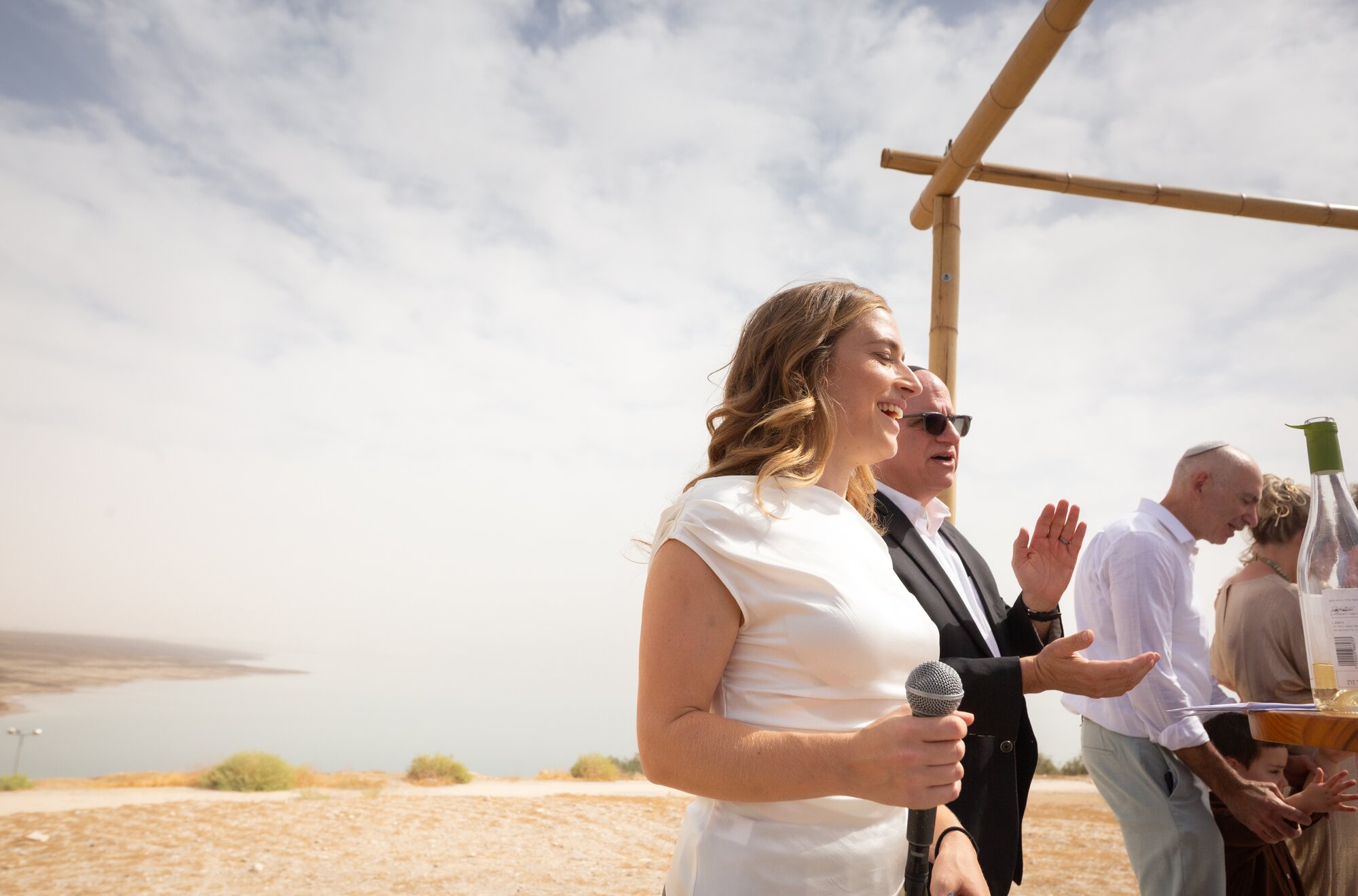 A guest speaking into a microphone beside the chuppah, the sea behind her