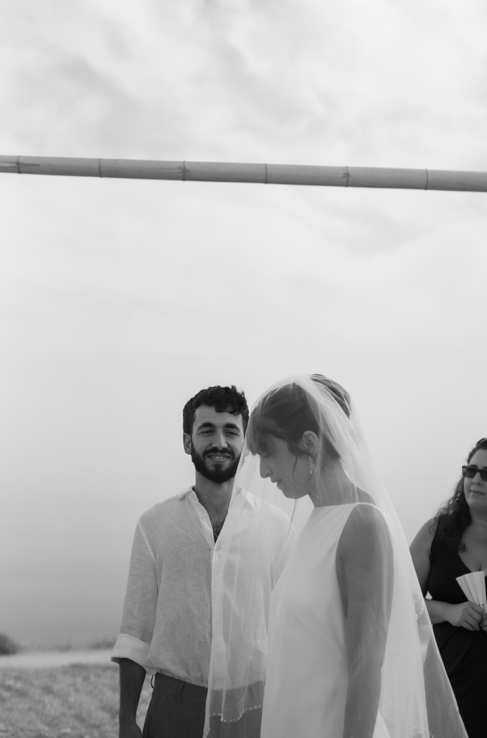 The groom smiling beside the veiled bride under the chuppah, black and white