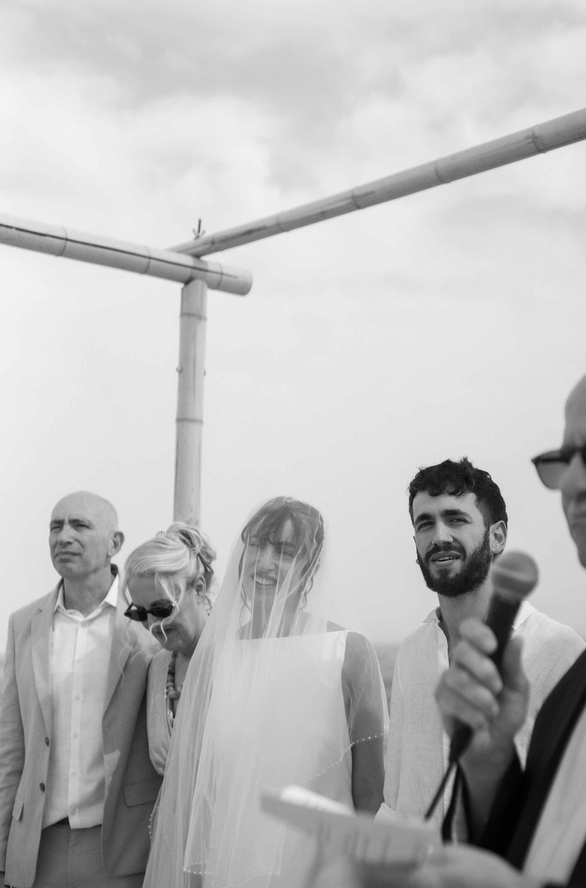 The groom speaking into a microphone beside the veiled bride and her parents, black and white