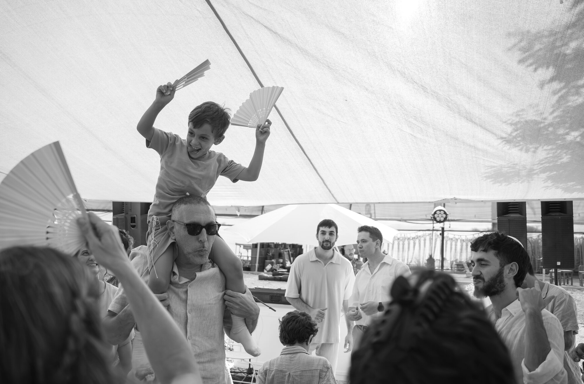 A child on a guest's shoulders waving a fan above the dancing crowd, black and white