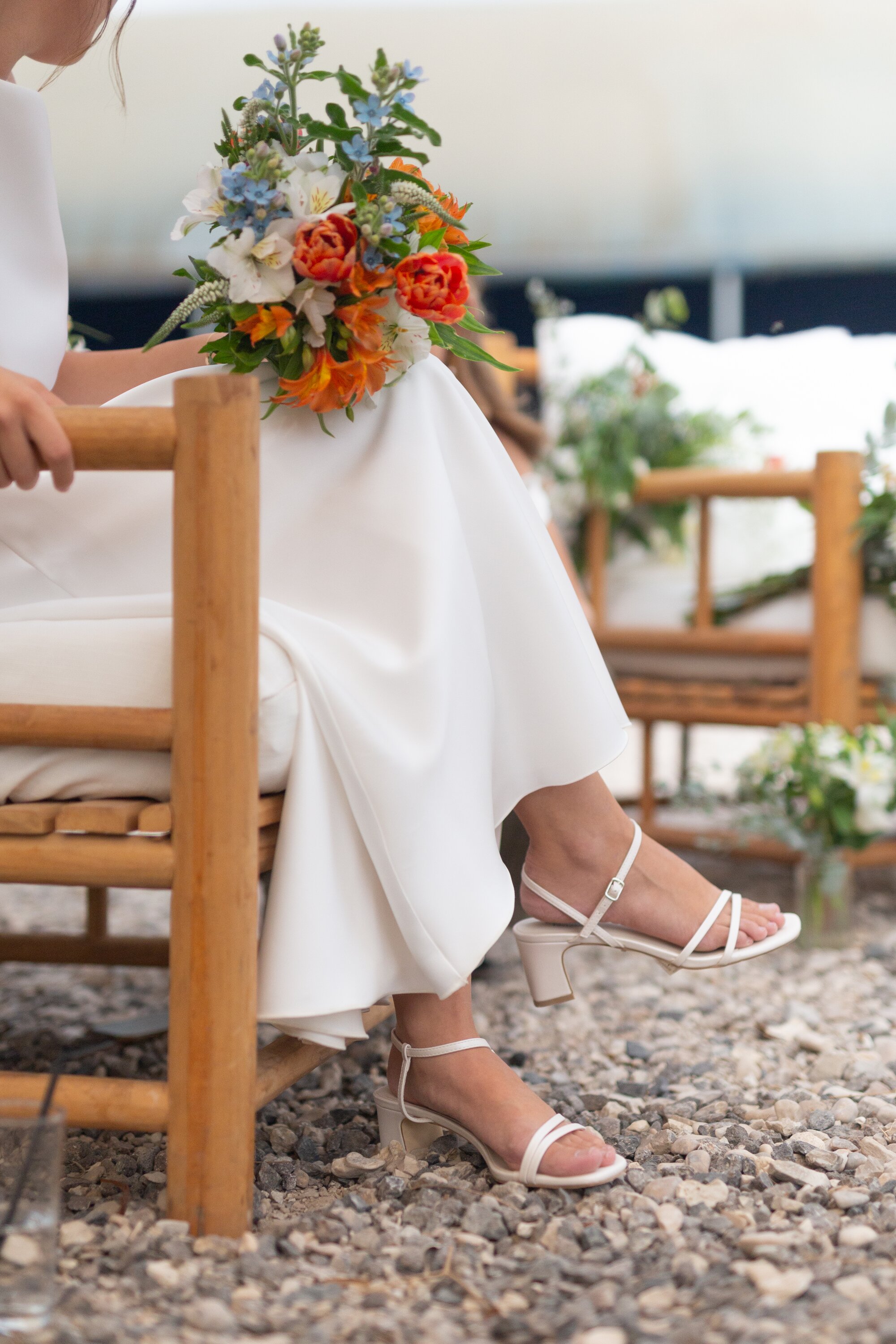 The bride seated on a wooden chair, her bouquet of orange and blue flowers on her lap