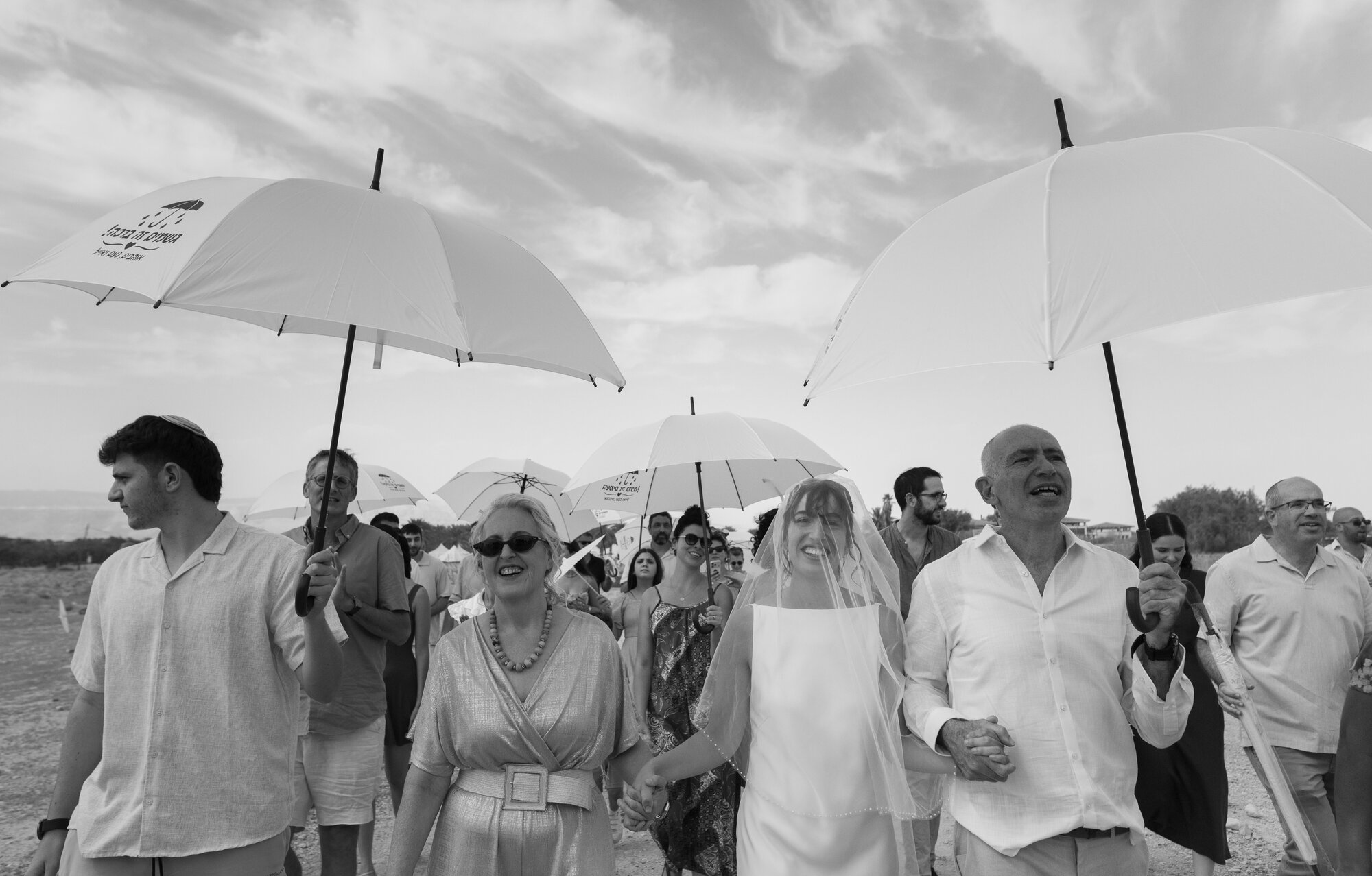 Guests walking beneath white umbrellas as the veiled bride is led to the chuppah between her parents, black and white