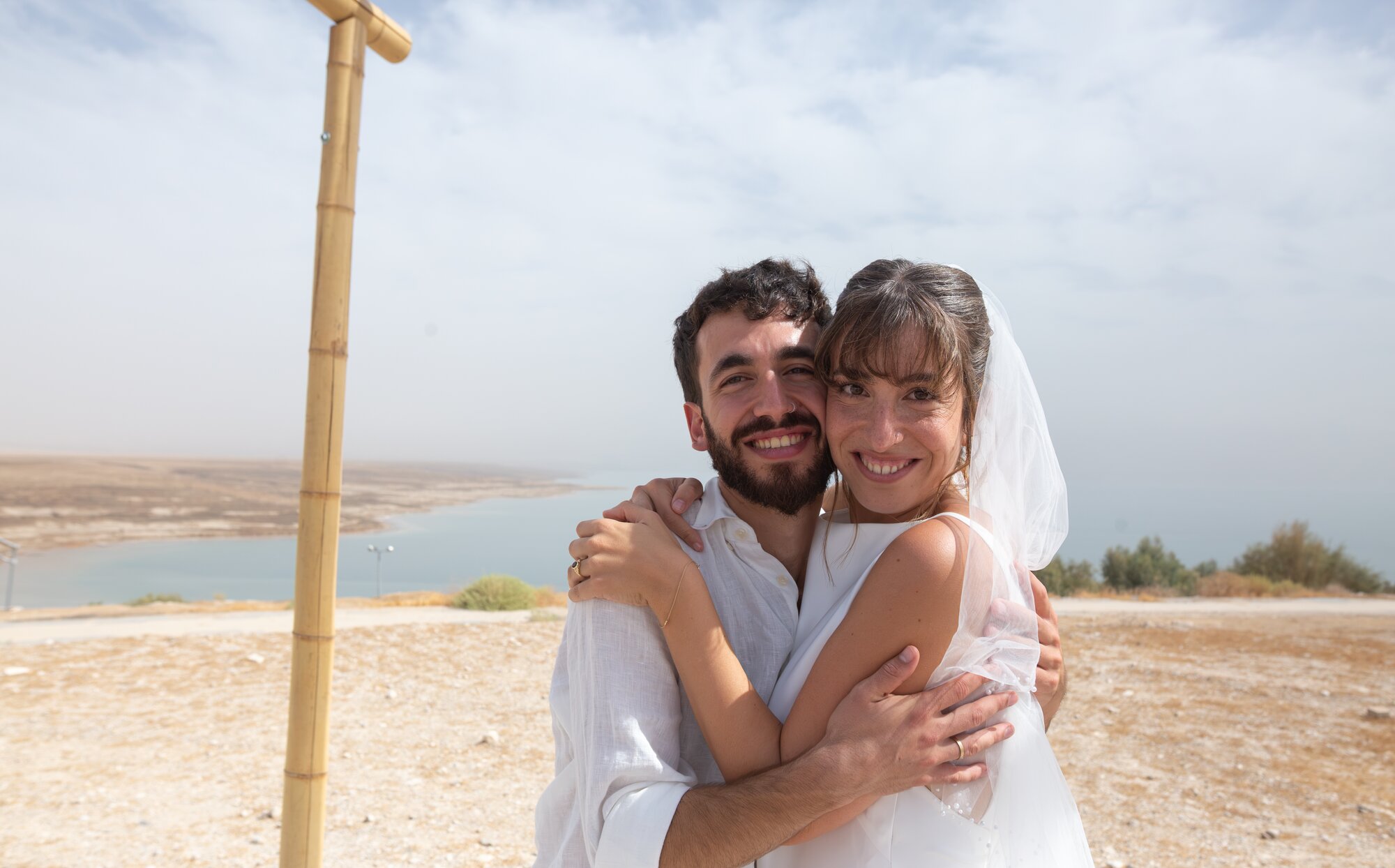 The bride and groom hugging and smiling, a bamboo pole beside them and the Dead Sea behind